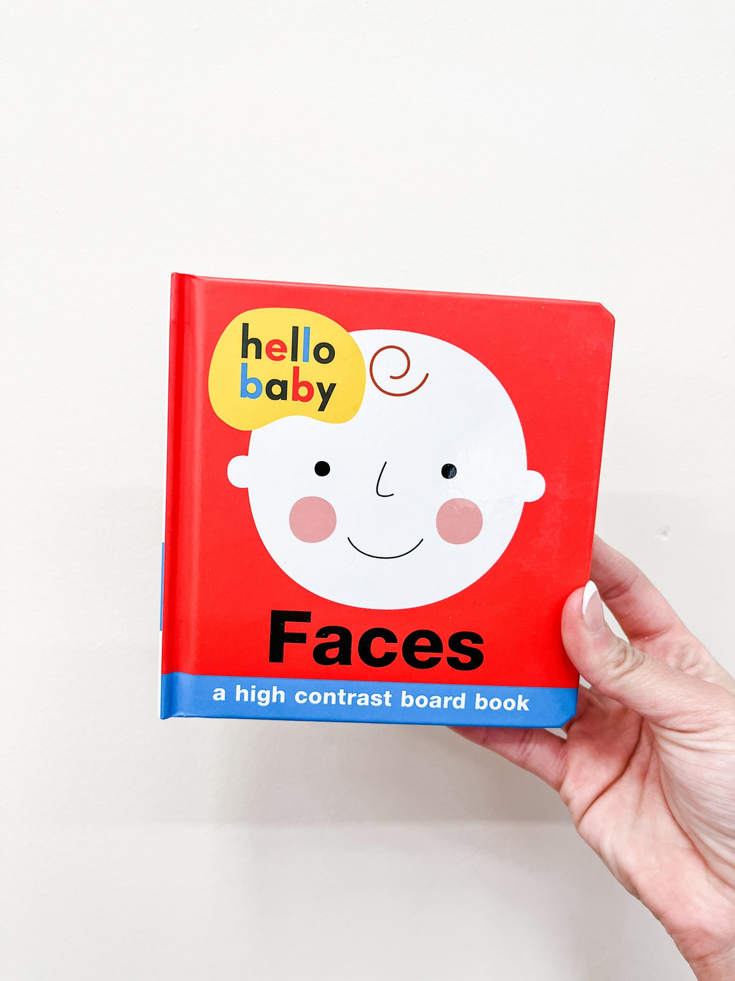 A hand holds the Hello Baby Faces - Board Book by Macmillan Publishers, with a red cover featuring a smiling baby’s face and the subtitle "a high contrast board book" against a plain white background.