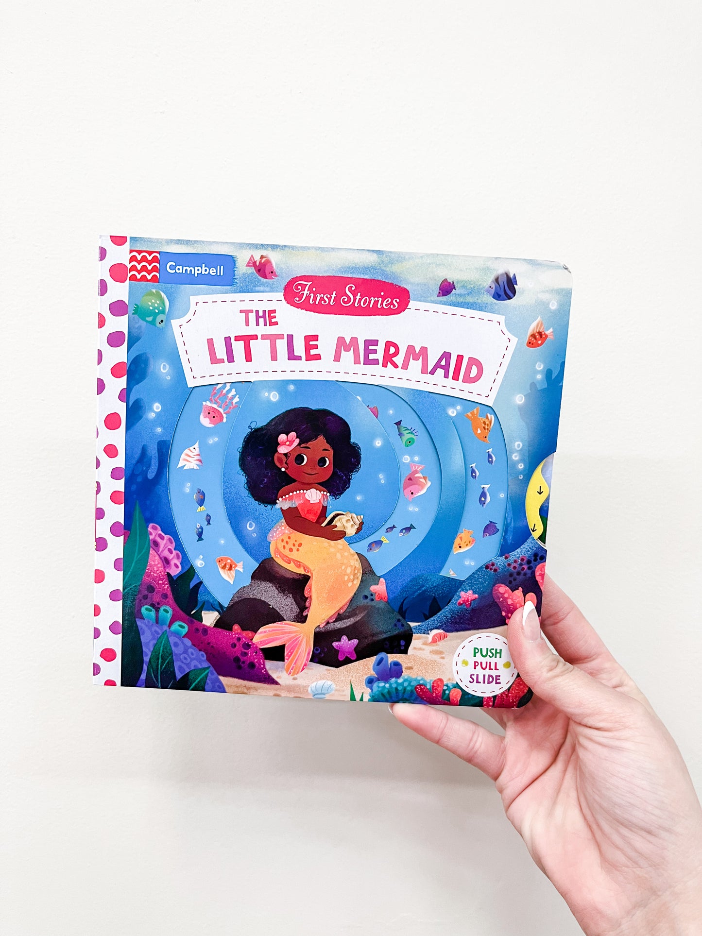 A hand holds the Little Mermaid Book by Macmillan Publishers. The cover shows a brown-skinned mermaid with dark curly hair sitting underwater, surrounded by fish and sea plants.
