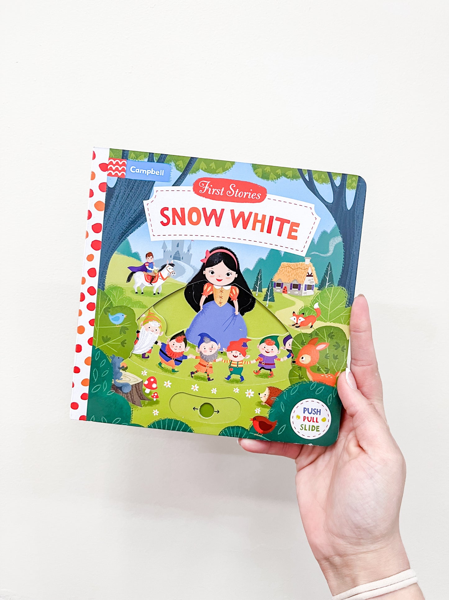 A hand holds the Snow White Book by Macmillan Publishers, a children's board book with colorful illustrations of Snow White, the seven dwarfs, forest animals, and a castle in the background.