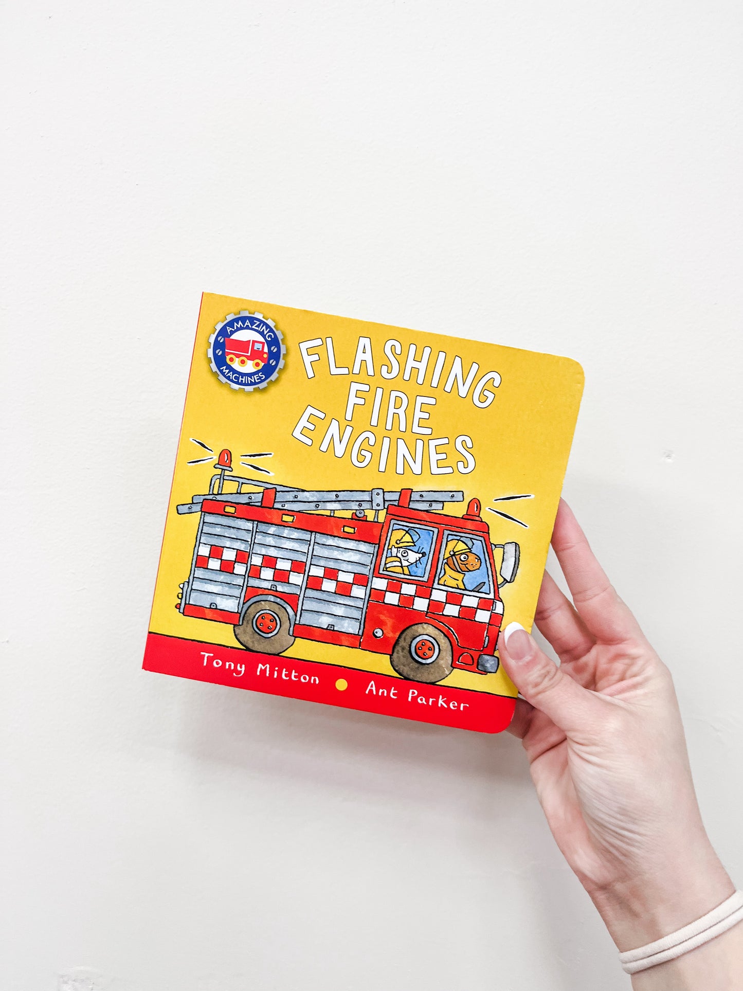 A hand holds the Macmillan Publishers' Flashing Fire Engines Book, featuring a yellow cover with a cartoon fire engine and two animal characters inside.