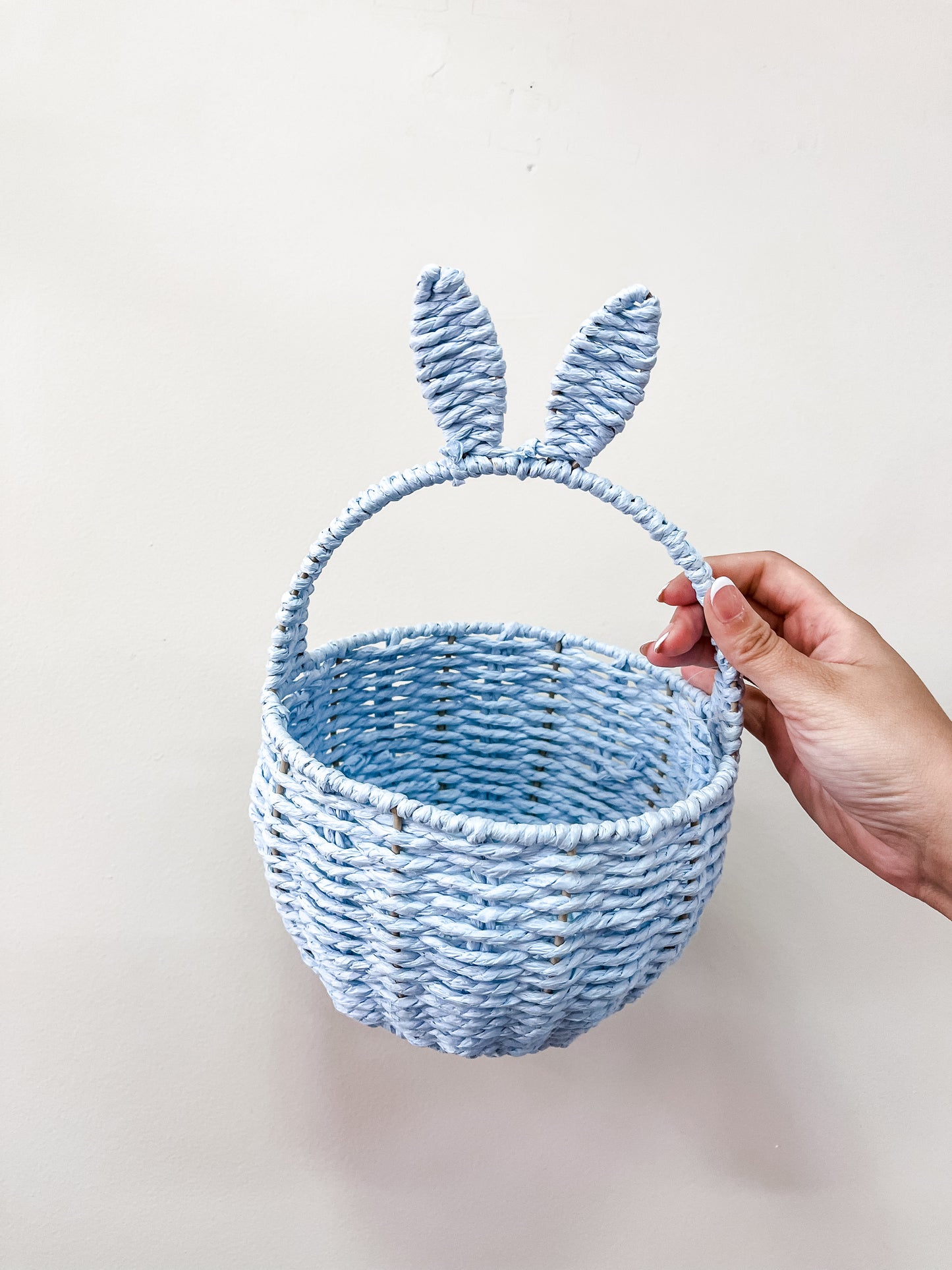 A hand holds the Capzone Pastel Bunny Ear Basket, a light blue woven basket with a handle adorned by two upright bunny ear decorations, set against a plain light background.