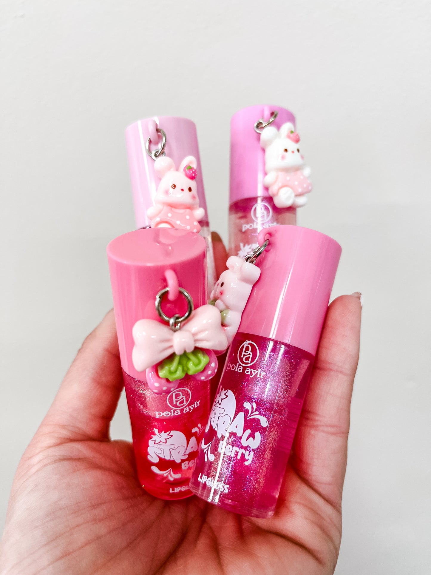 A hand holds four Capzone Strawberry Charm Shimmery Lip Gloss tubes, each featuring a glittery "strawberry" label, a bunny charm, and a bow keychain on the lid, all set against a plain white background.