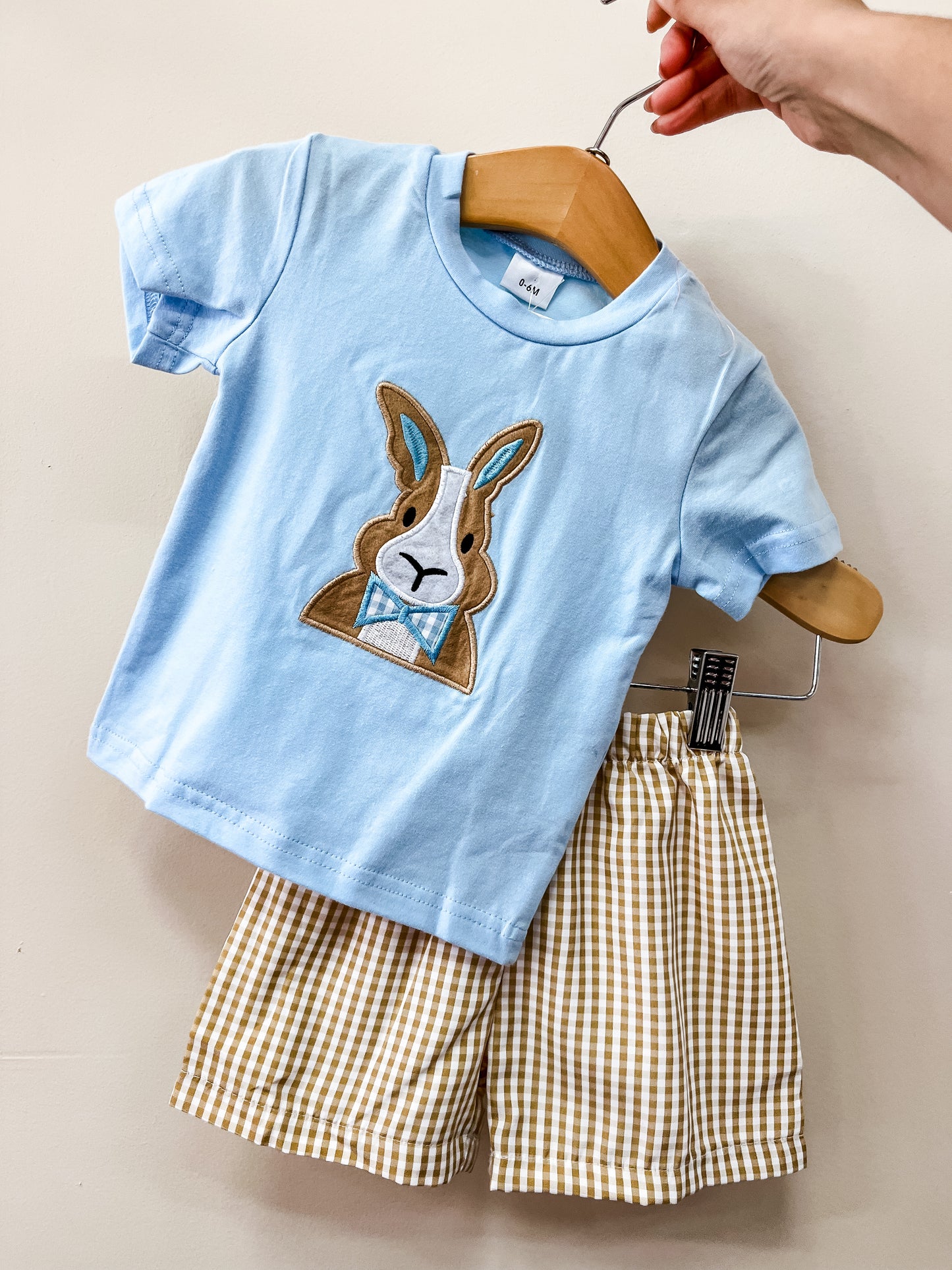 A hand holds the Tiny Tot Bundles Bunny Bowtie Short Set—a child's light blue t-shirt with a rabbit appliqué and bow tie, paired with beige and white checkered shorts.