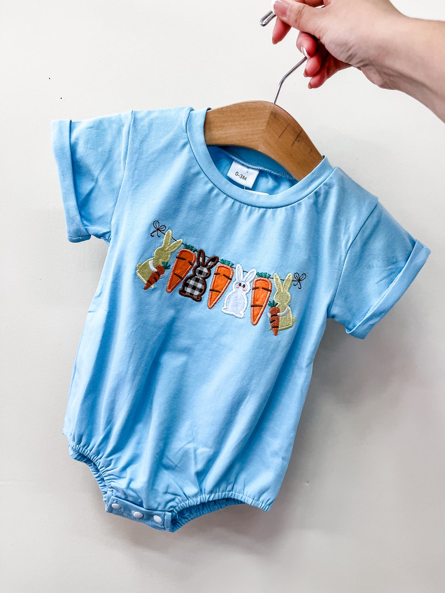 The Hoppy Carrot Crew Bubble by Tiny Tot Bundles is a light blue baby romper with embroidered carrots and bunny rabbits across the chest, displayed on a wooden hanger against a plain white background.