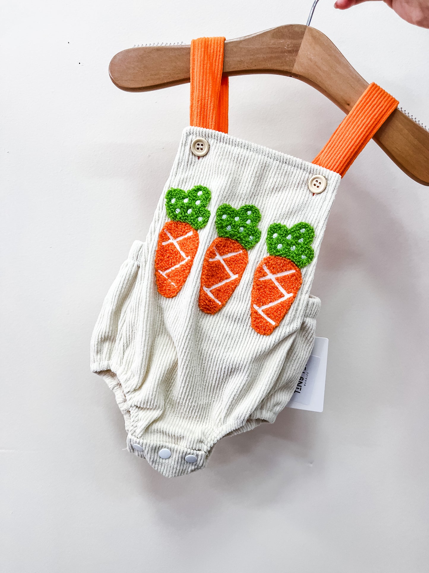 The Bunny Cord Overalls by Tiny Tot Bundles are cream-colored baby rompers with orange suspenders and three embroidered orange and green carrots on the front, shown hanging on a wooden hanger against a white background.