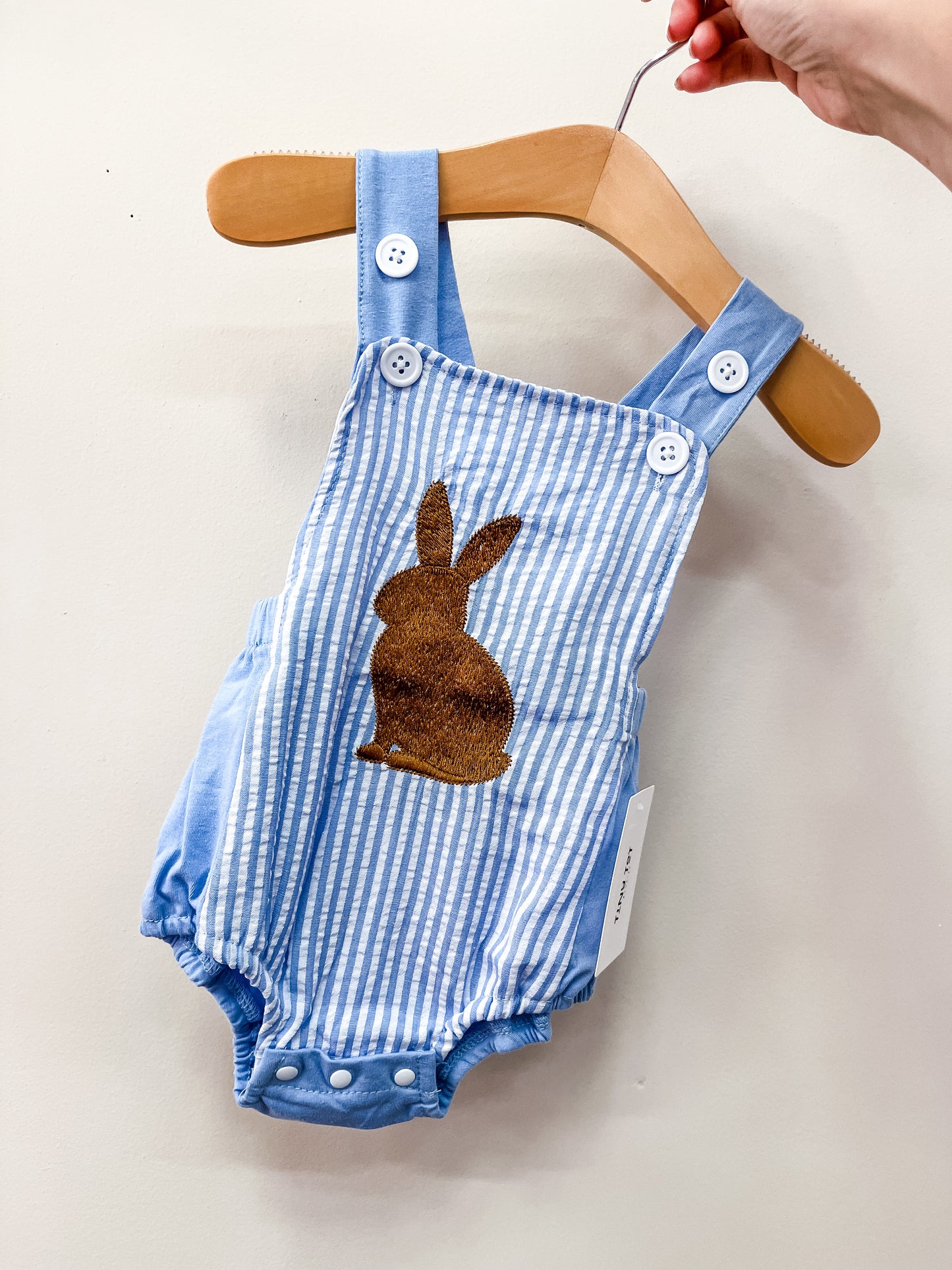 The Tiny Tot Bundles Blue Bunny Overalls feature blue and white stripes, white buttons, and a brown bunny silhouette on the front, shown on a wooden hanger held by hand against a white background.