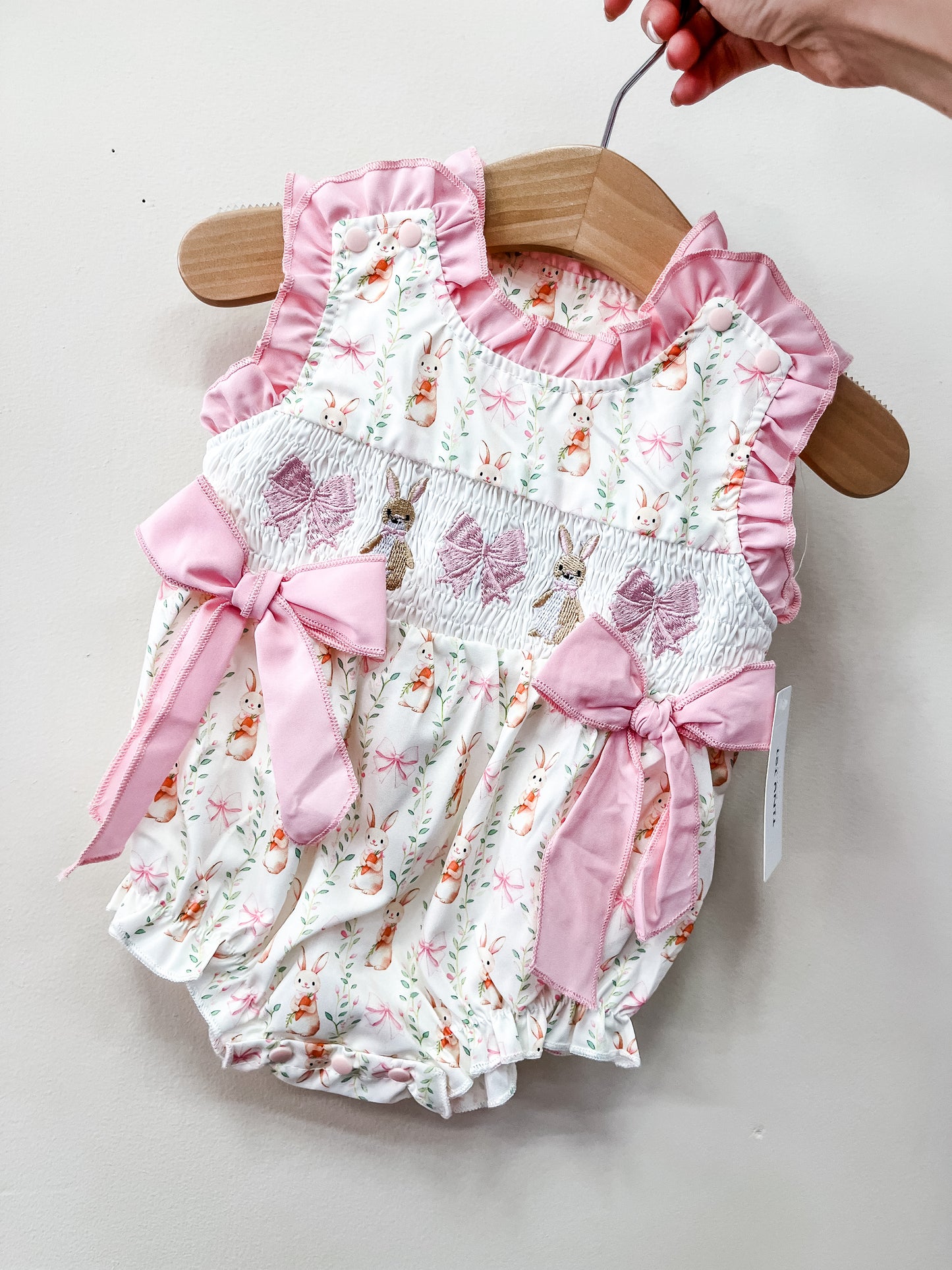 The Blushing Bunny Bow Bubble by Tiny Tot Bundles features a pink and white bunny pattern, pink ruffled trim, and two pink bows. Displayed on a wooden hanger held by hand against a plain background.