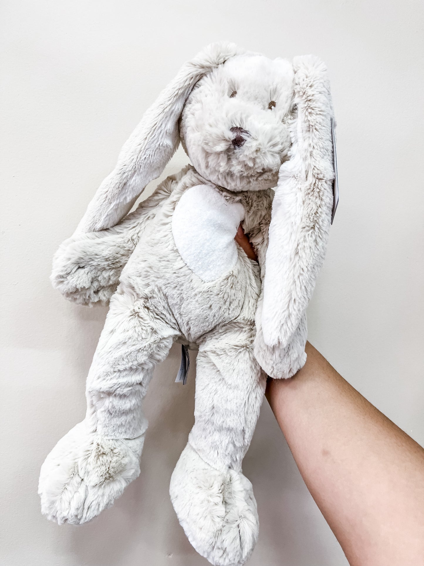 A hand holds up the Tiny Tot Bundles Grey Heart Bunny Plush, a soft, light grey stuffed bunny with long floppy ears and stitched facial features, set against a plain off-white background.