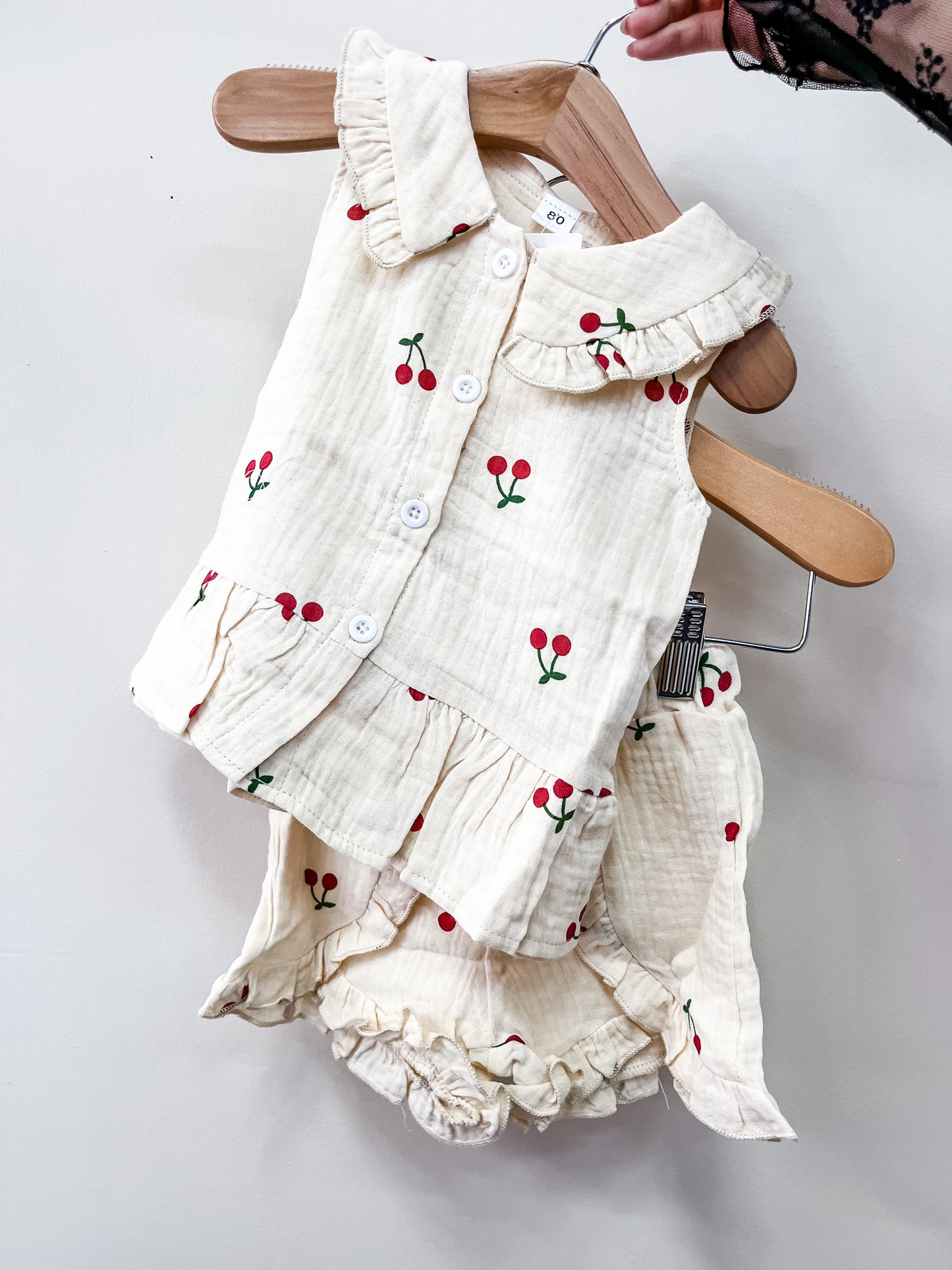 The Cherry Cream Ruffle Set by Tiny Tot Bundles features a cream-colored sleeveless ruffled top and matching bloomers with a red cherry print, displayed on wooden hangers against a white background.