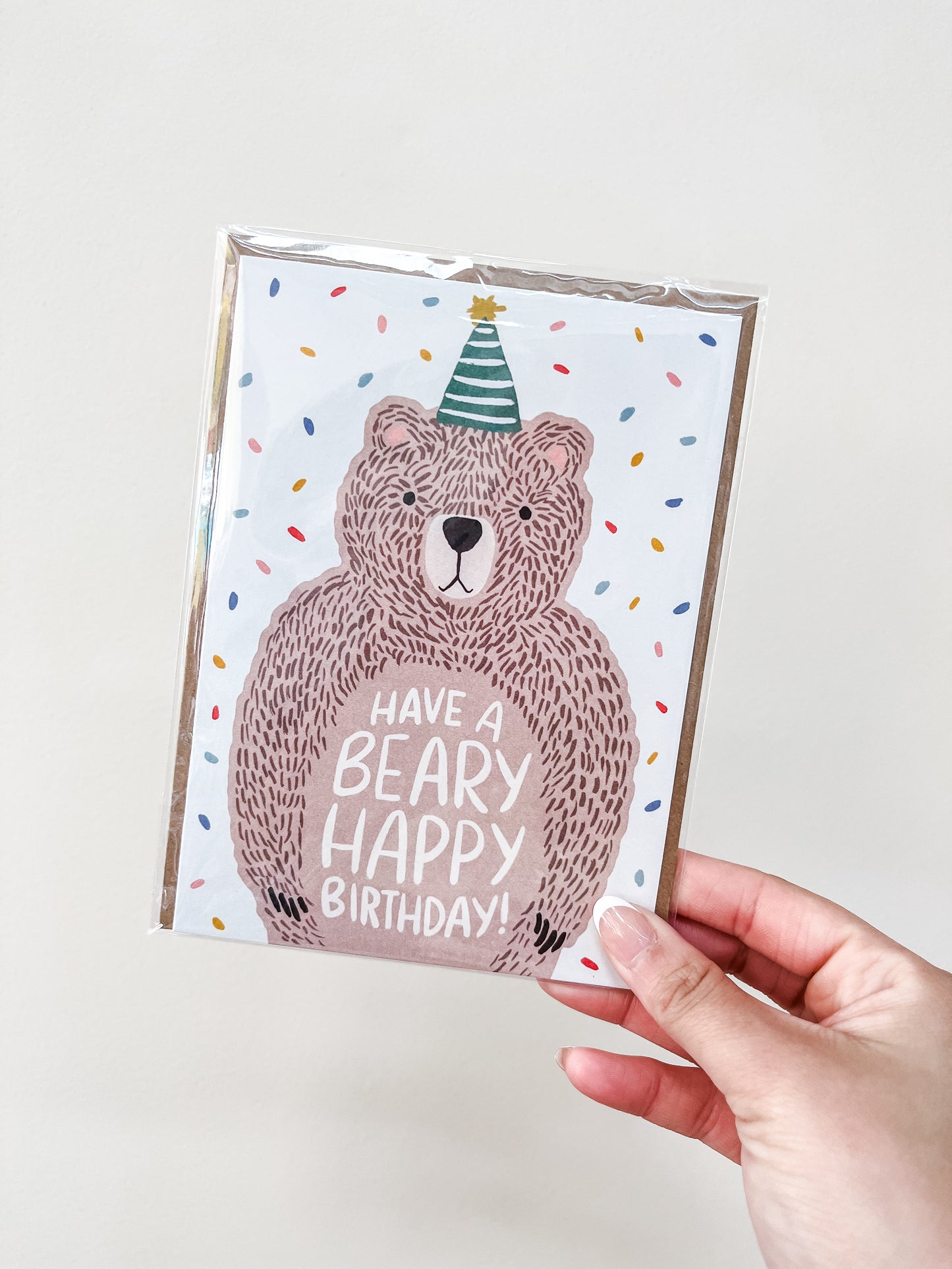 A hand holds the "Bear - Birthday Card" by love Light Paper, featuring a cartoon bear in a party hat with confetti. The card says “Have a beary happy birthday!” in playful lettering.