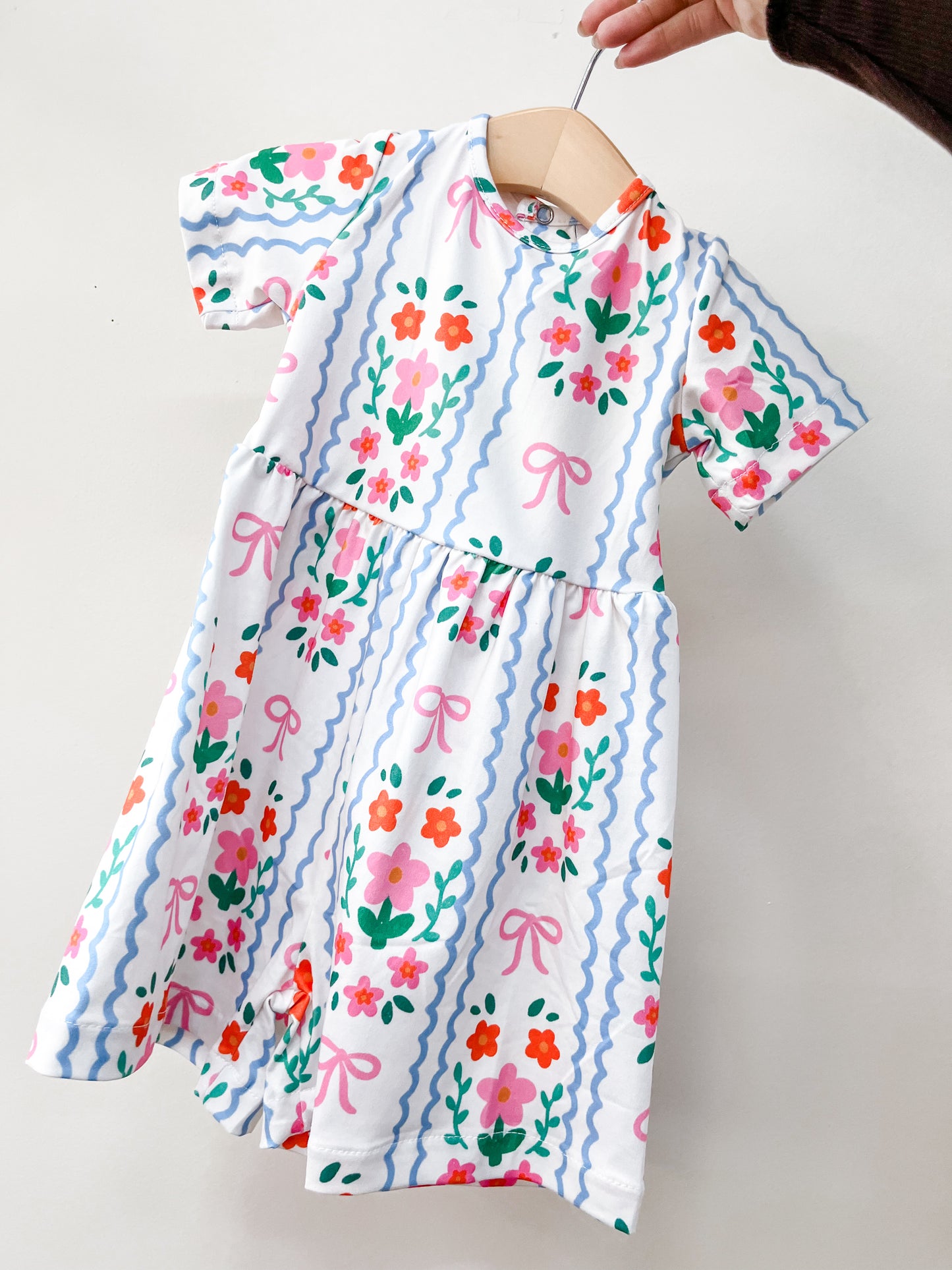 A hand holds up the Tiny Tot Bundles Ribbon Garden Romper, a short-sleeved white baby romper featuring colorful flowers, pink bows, blue wavy stripes, a gathered waist, and a vibrant design.