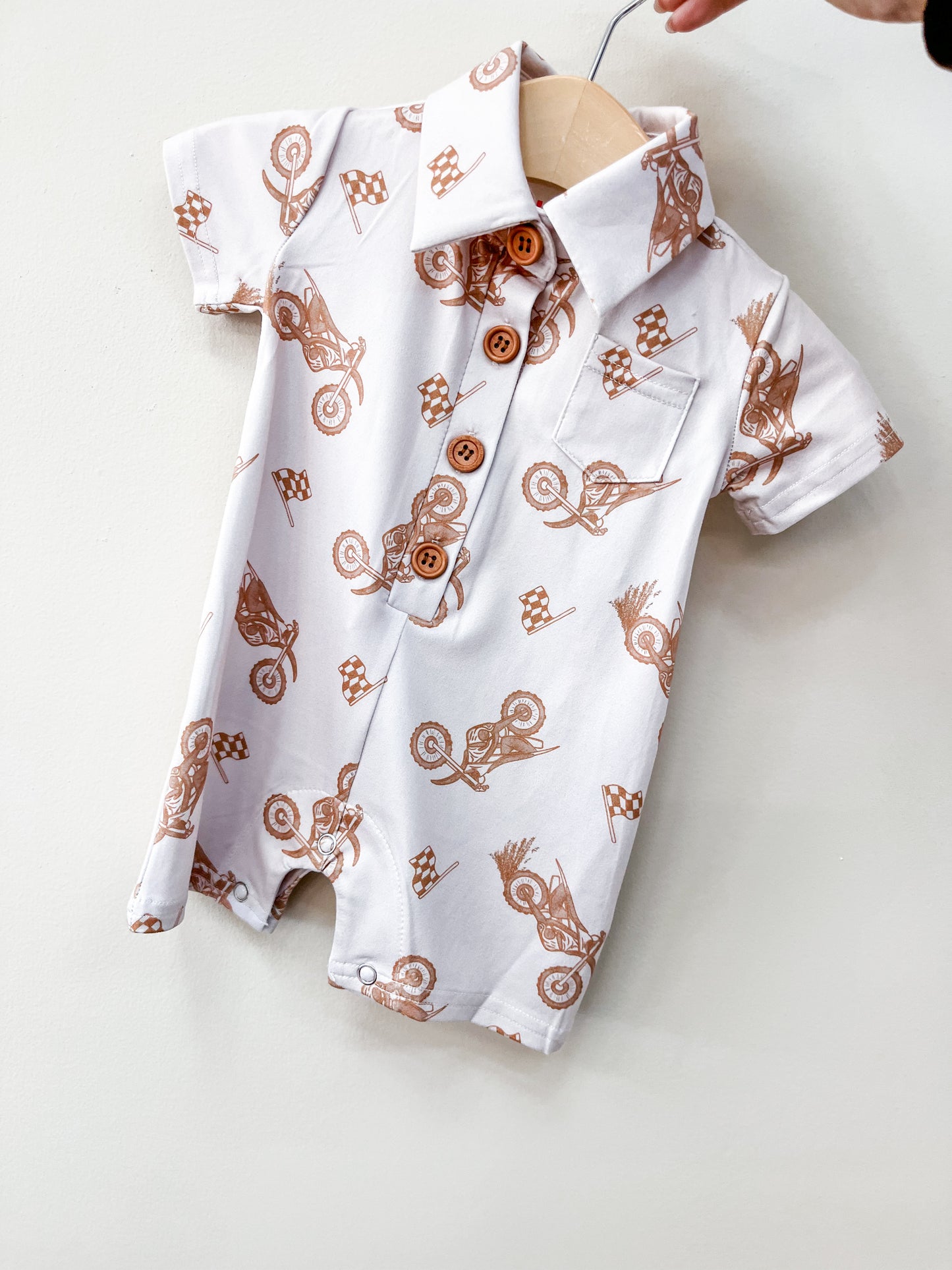 The Moto Ride Collard Romper by Tiny Tot Bundles is a short-sleeved white baby romper with brown motocross bike and checkered flag prints, wooden buttons, and a front pocket, displayed on a wooden hanger against a light background.