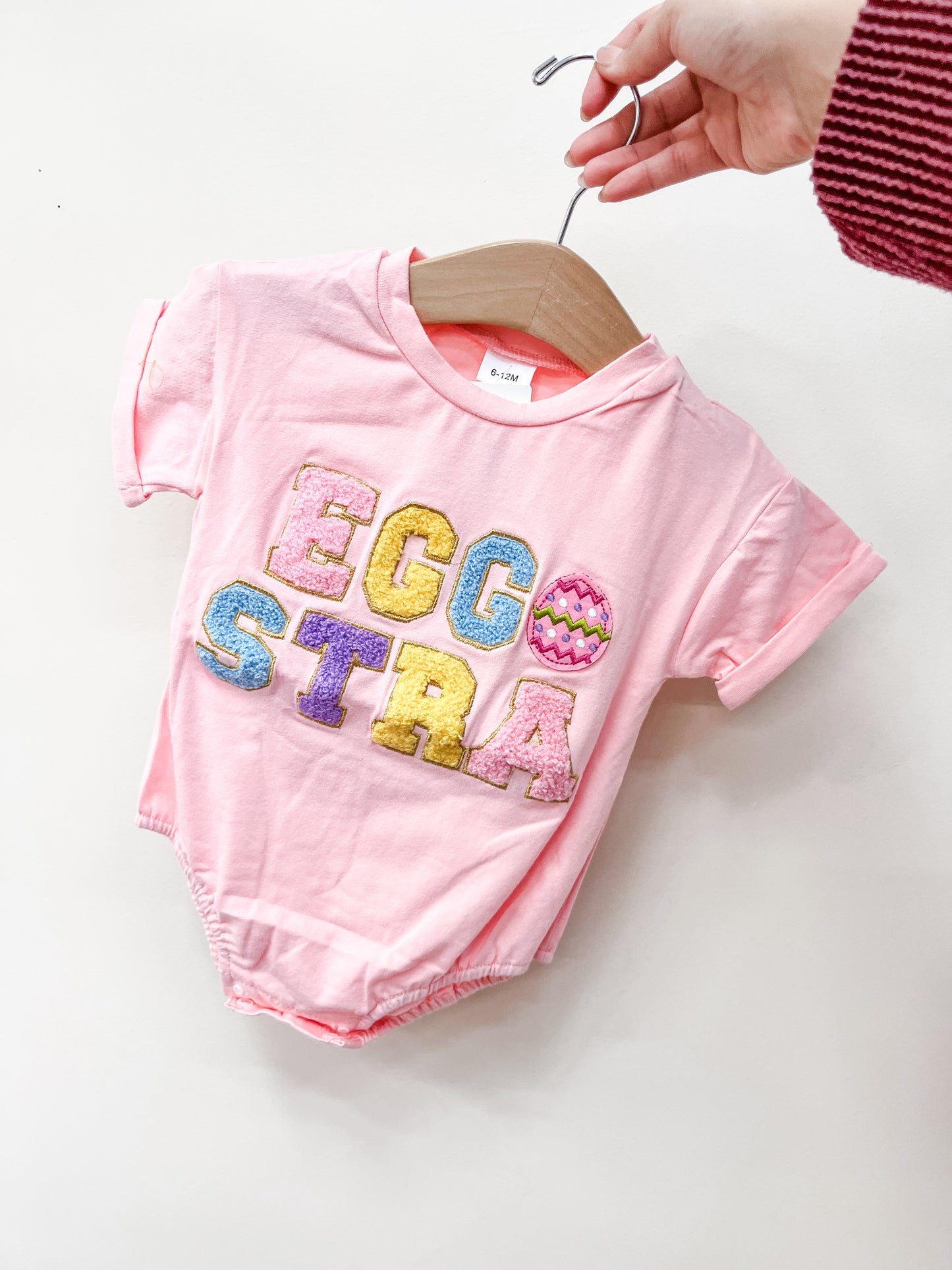 A hand holds the Tiny Tot Bundles Egg Hunt Bubble, a pink baby onesie on a wooden hanger. The front displays colorful, textured letters spelling EGGSTRA, with an Easter egg replacing the letter O.