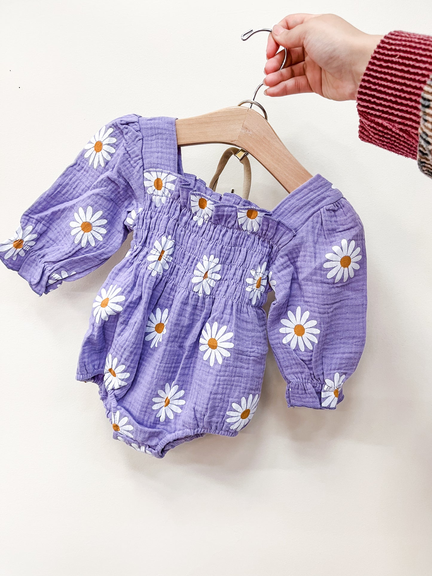 A hand holds a hanger with the Tiny Tot Bundles Lavender Daisy Romper, a purple baby romper featuring white daisies with yellow centers, against a plain light background.