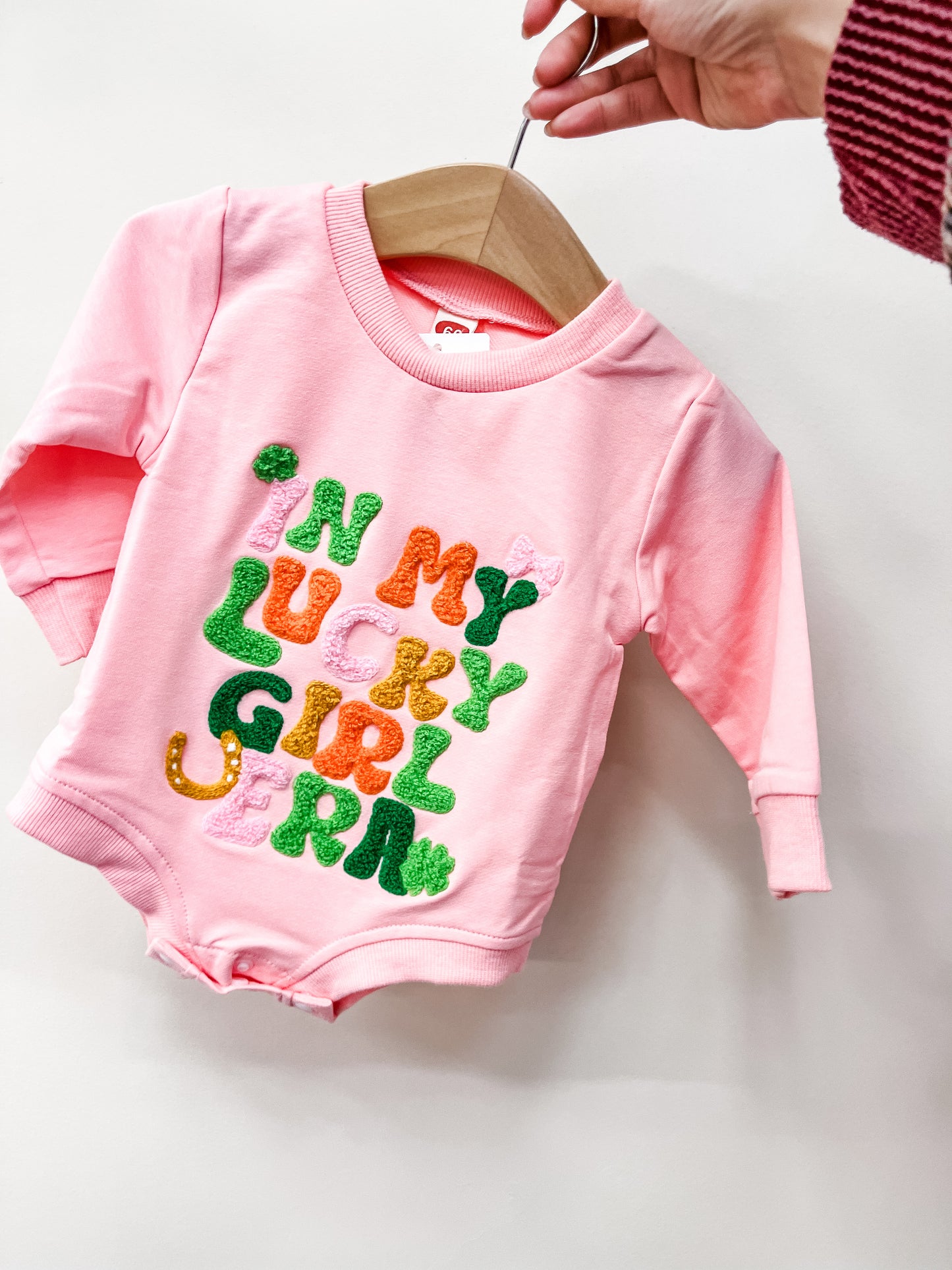 A hand holds up the Pink Lucky Girl Bubble by Tiny Tot Bundles—a pink baby romper with colorful, textured letters spelling IM MY LUCKY GIRL GERM on the front.