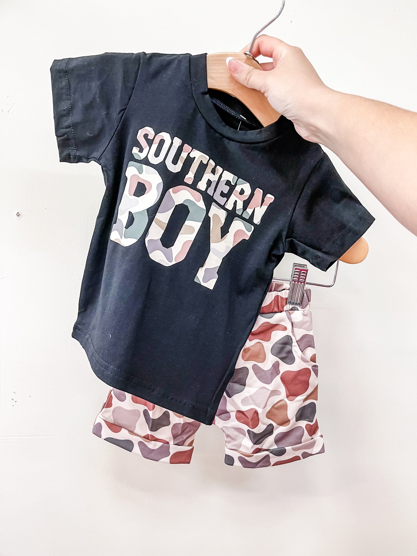 A hand holds up the Tiny Tot Bundles Southern Boy Short Set, which includes a black t-shirt with "Southern Boy" in camouflage letters and multicolor heart-patterned shorts.