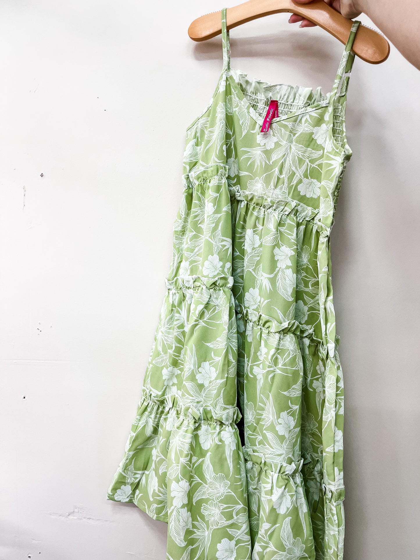 A hand holds the Key Lime Garden Dress by Cutie Patootie—a green floral tiered sundress with thin straps and ruffle details—displayed on a wooden hanger against a plain white wall.