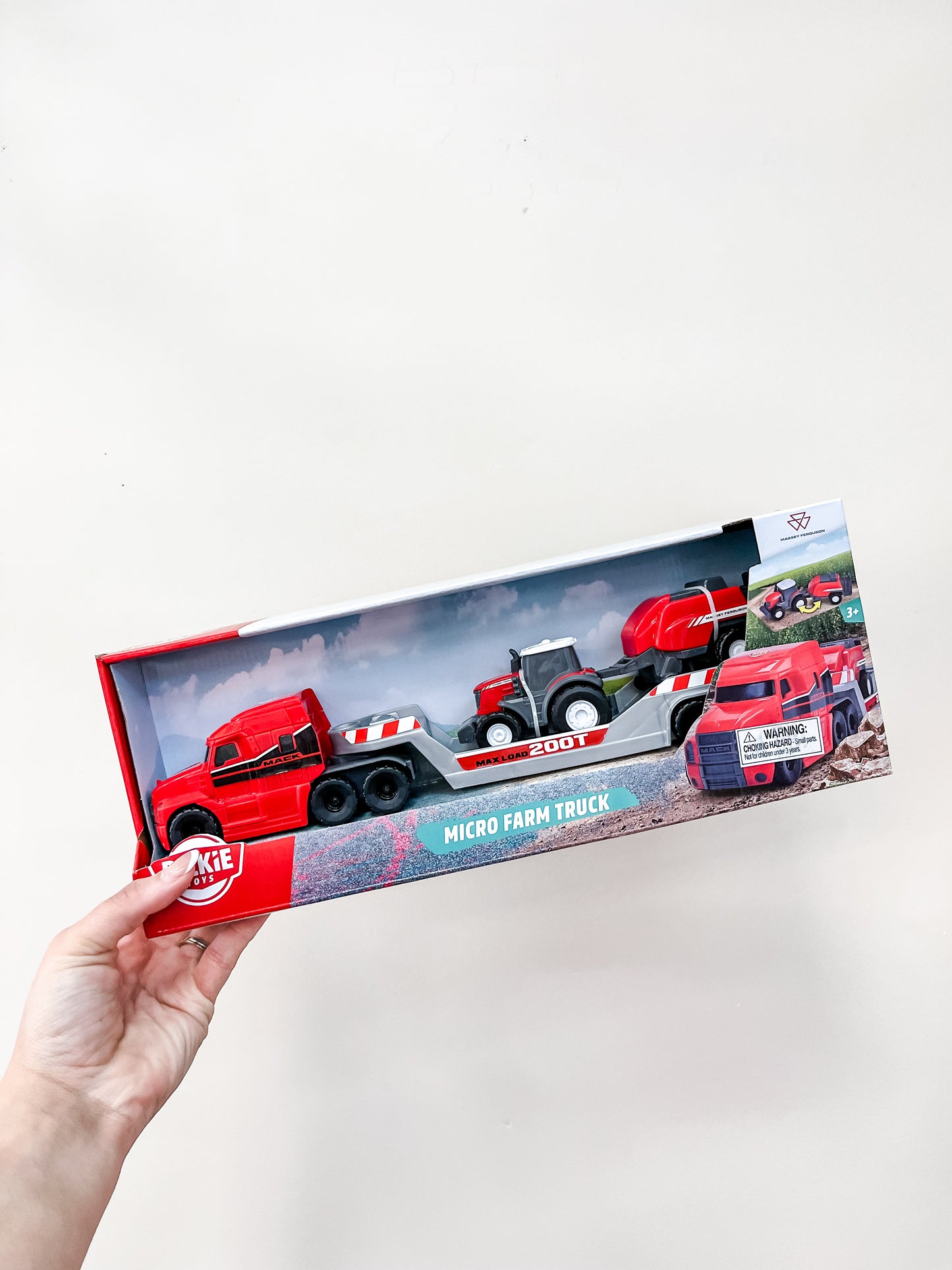A hand holds a Toy Smith Micro Farm Truck Assortment boxed set, featuring a red semi-truck, tractor, and trailer in clear packaging against a plain white background.