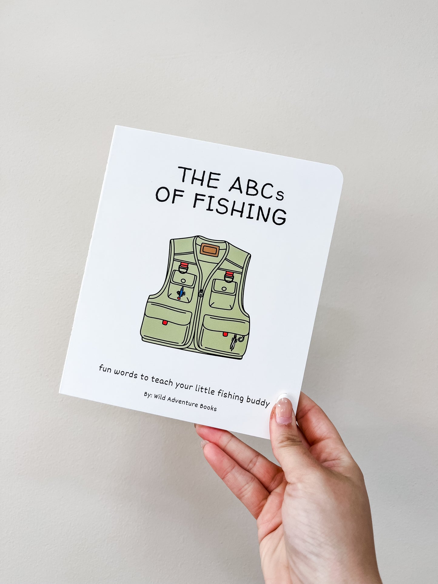 A hand holds the Abc's of Fishing Book by Wild Adventure Books, featuring a green fishing vest illustration on the cover, set against a plain light background.