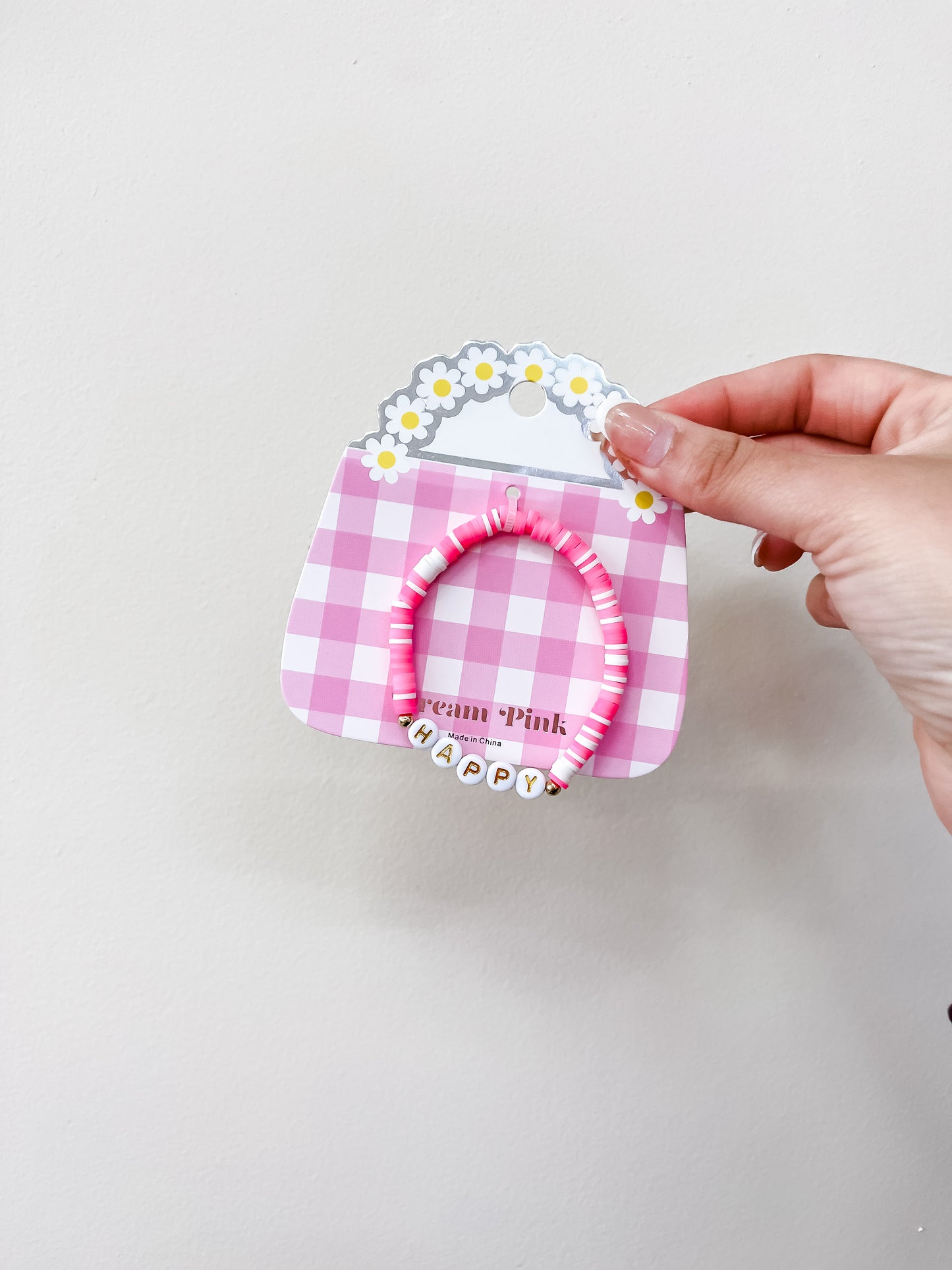 A hand holds the funteze Happy Words Bracelet, featuring pink and white beads and letter beads spelling HAPPY, displayed on a pink gingham card with daisy decorations.