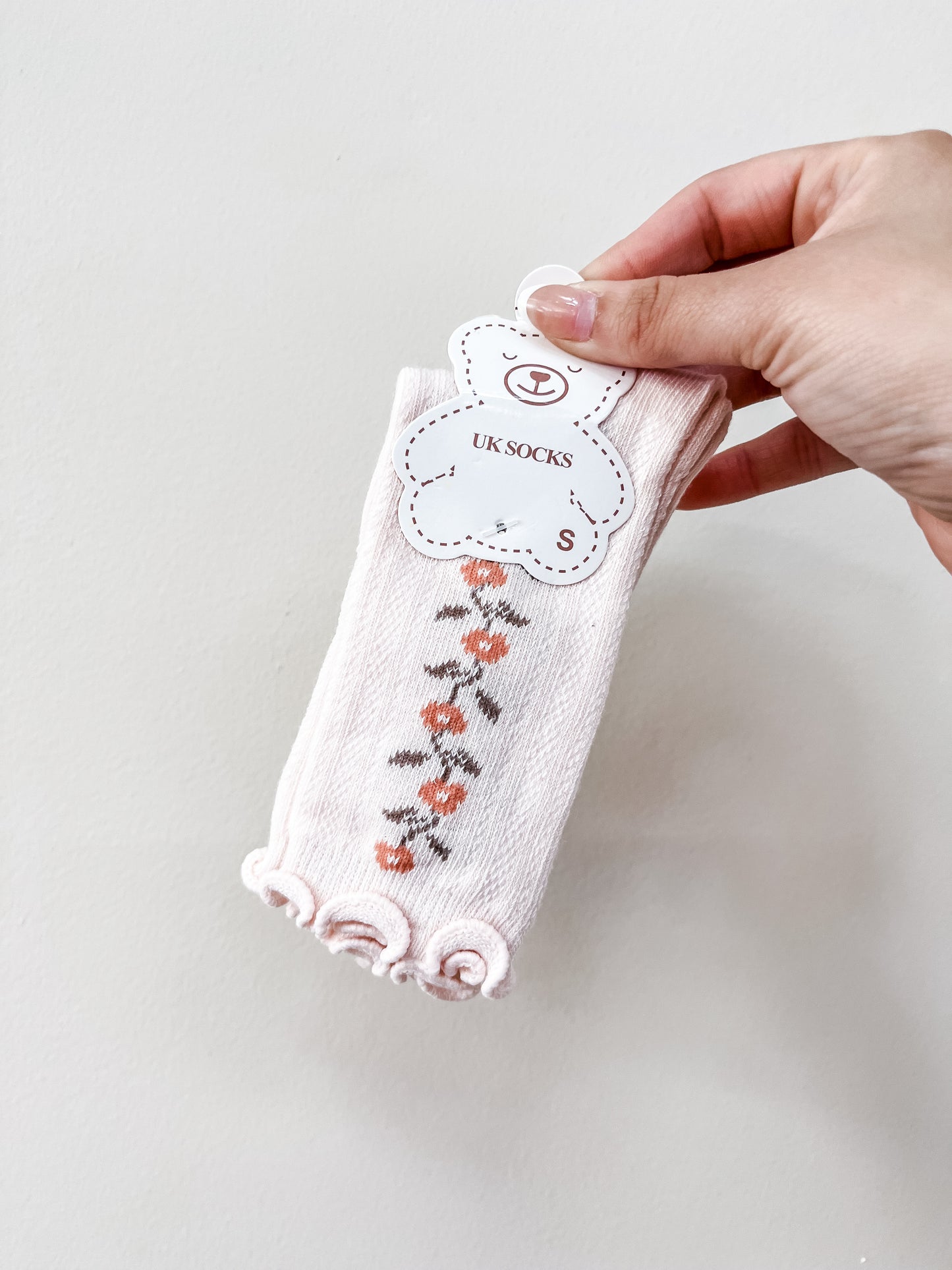 A hand holds a pair of Tiny Tot Bundles Pink Petal Socks featuring a floral pattern and frilled edges, labeled with a bear-shaped tag.
