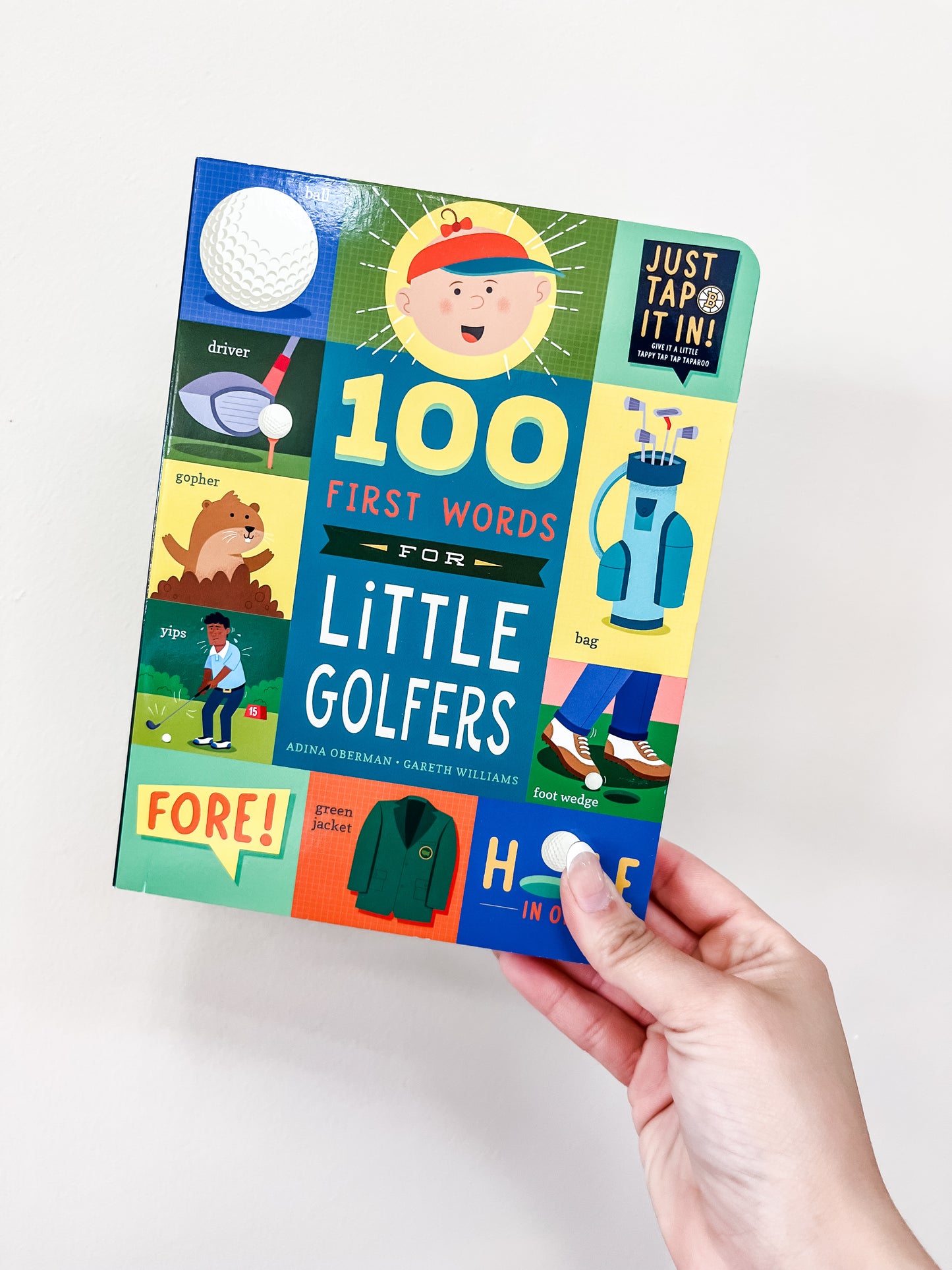 A hand holds the familuis "100 First Words For Little Golfers Book," a colorful children’s book with playful golf-themed illustrations like a golf ball, golf bag, and cartoon golfer on a white background.