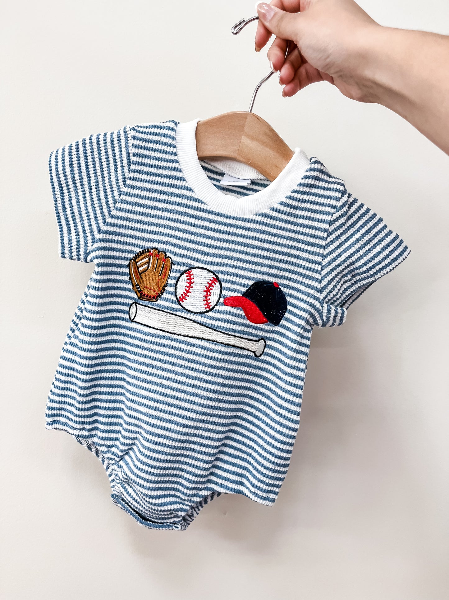 A hand holds the Tiny Tot Bundles "Little Slugger Bubble"—a blue-and-white striped baby romper adorned with embroidered baseball glove, ball, bat, and cap on the front.