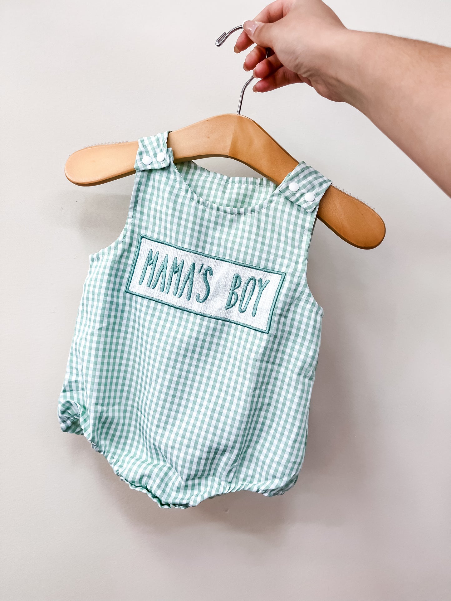 A hand holds a wooden hanger displaying the Tiny Tot Bundles Mama's Boy Gingham Bubble, a green and white checkered baby romper with buttoned straps and "MAMA'S BOY" embroidered on the front.