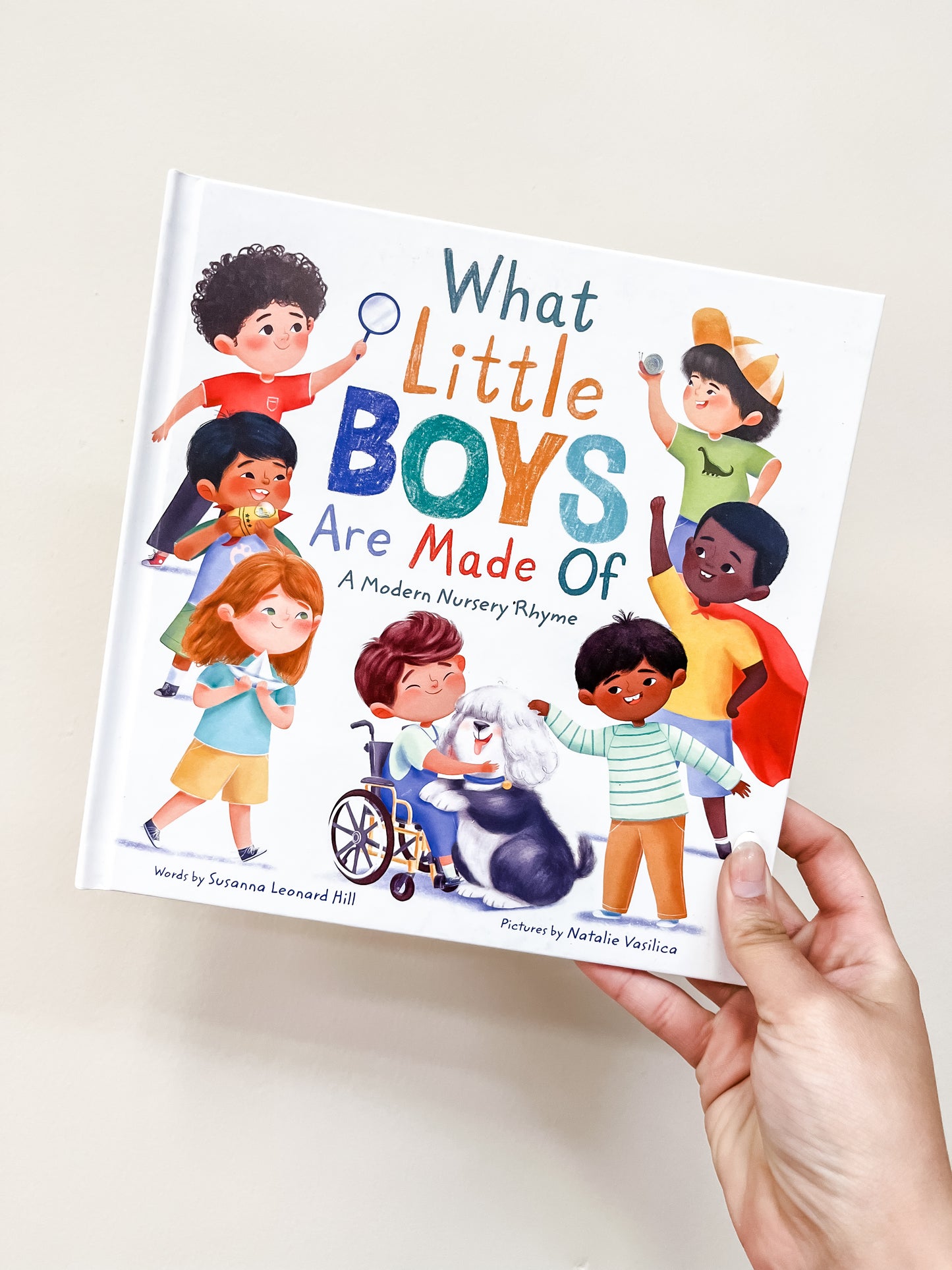 A hand holds the Sourcebooks "What Little Boys Are Made of Book," featuring a cover with diverse boys in playful costumes, smiling and interacting.