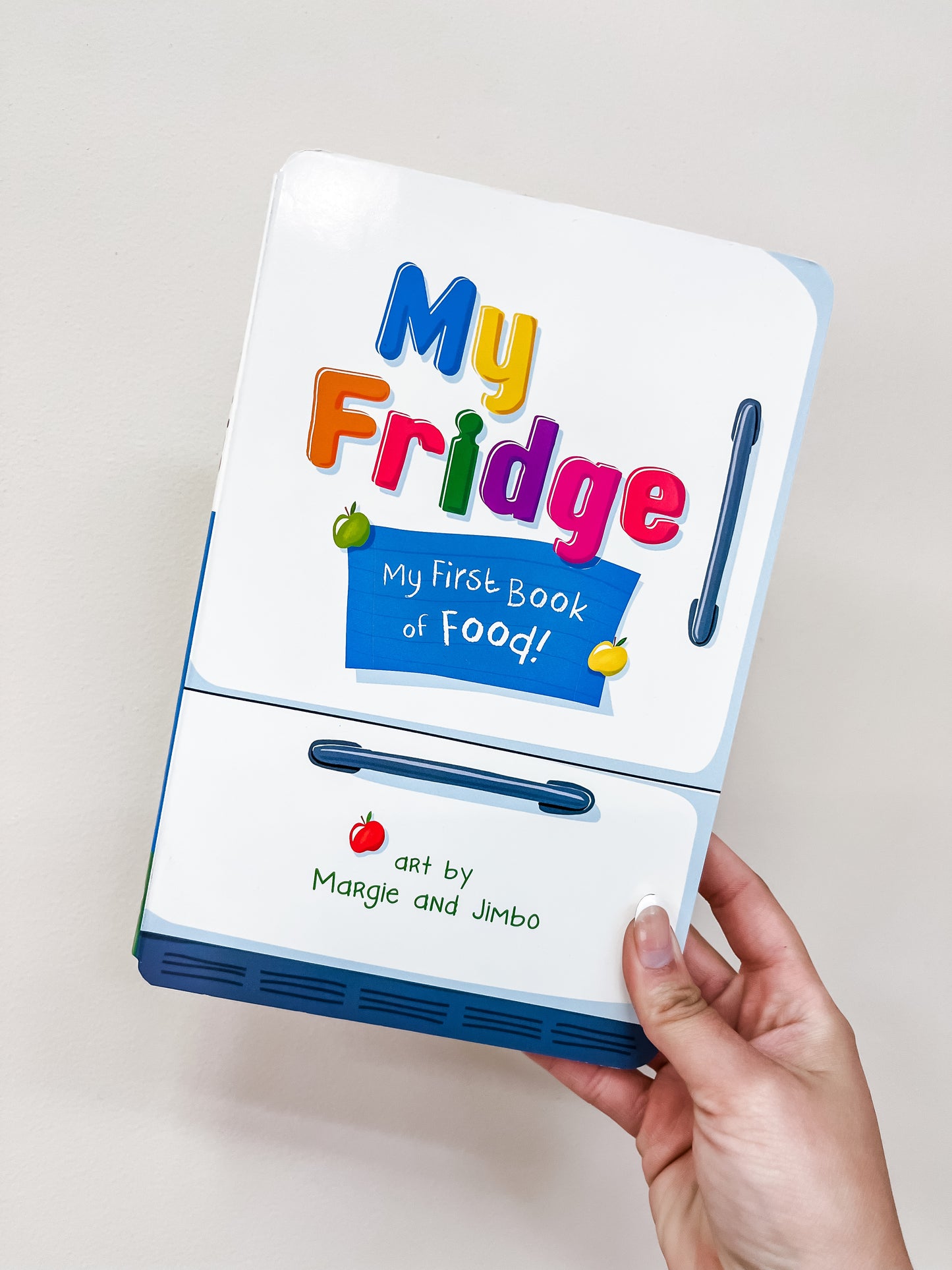 A hand holds the Sourcebooks My Fridge Book, a children's book with colorful lettering and food illustrations on a cover designed like a refrigerator.