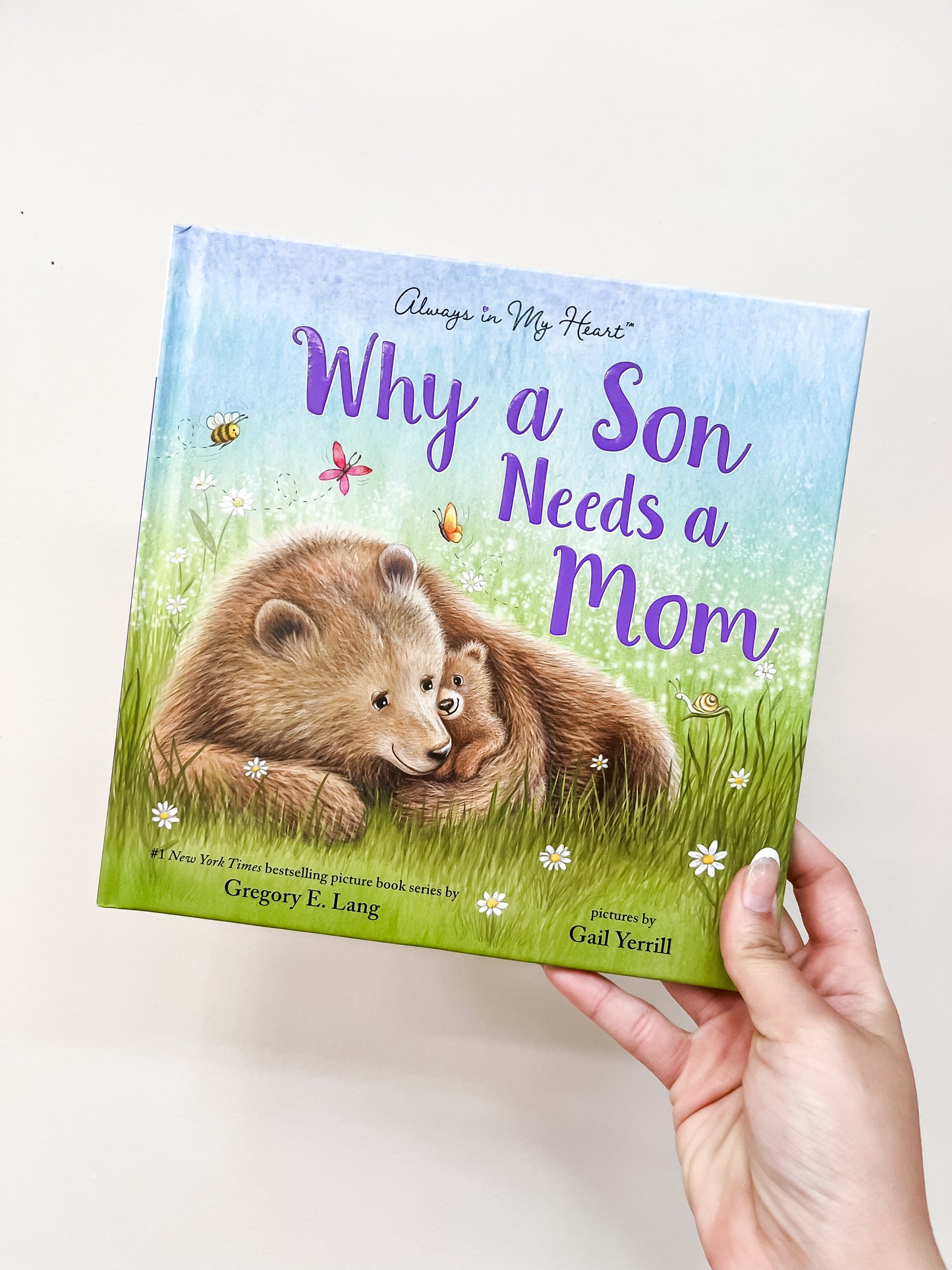 A hand holds the Why A Son Needs A Mom Book by sour, which has an illustration of a mother and son bear cuddling in a grassy field with butterflies and flowers.