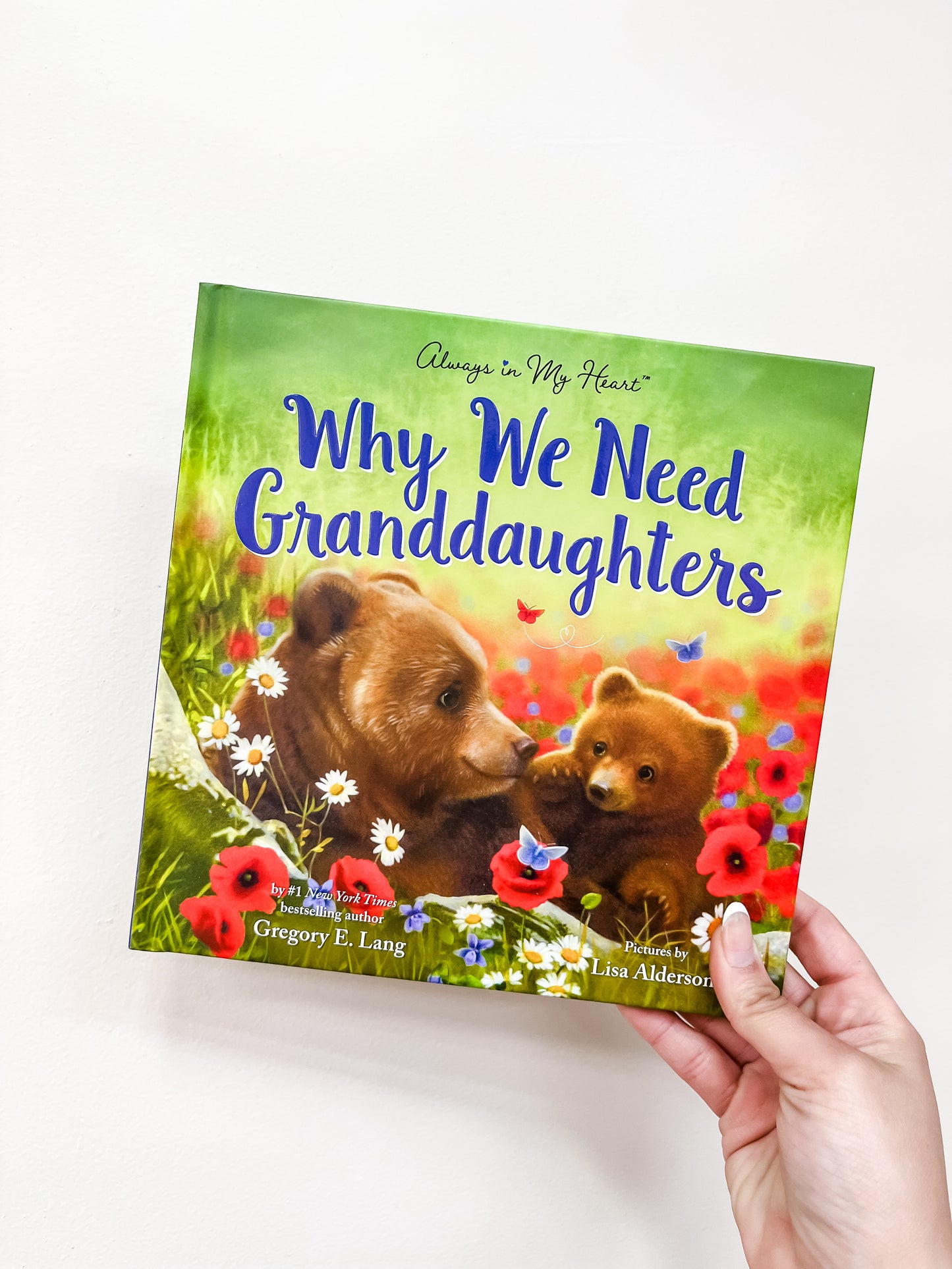 A hand holds the Why We Need Granddaughters Book by sour, featuring a cover illustration of two brown bears surrounded by red flowers.