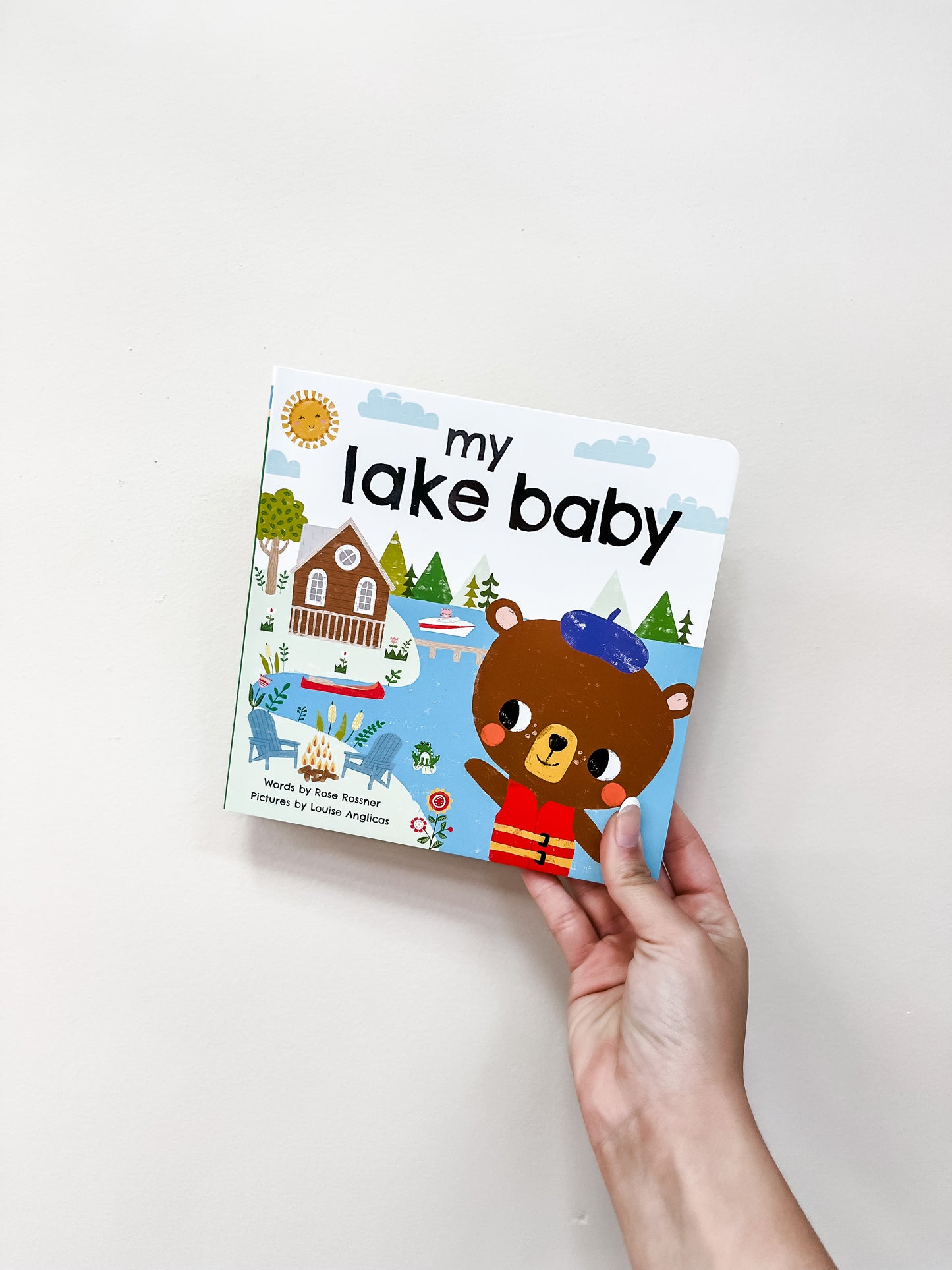 A hand holds the "My Lake Baby Book" by Sourcebooks, a children's board book with an illustrated bear in a blue hat and red outfit, plus a lakeside cabin scene with trees, chairs, and a canoe on the cover.