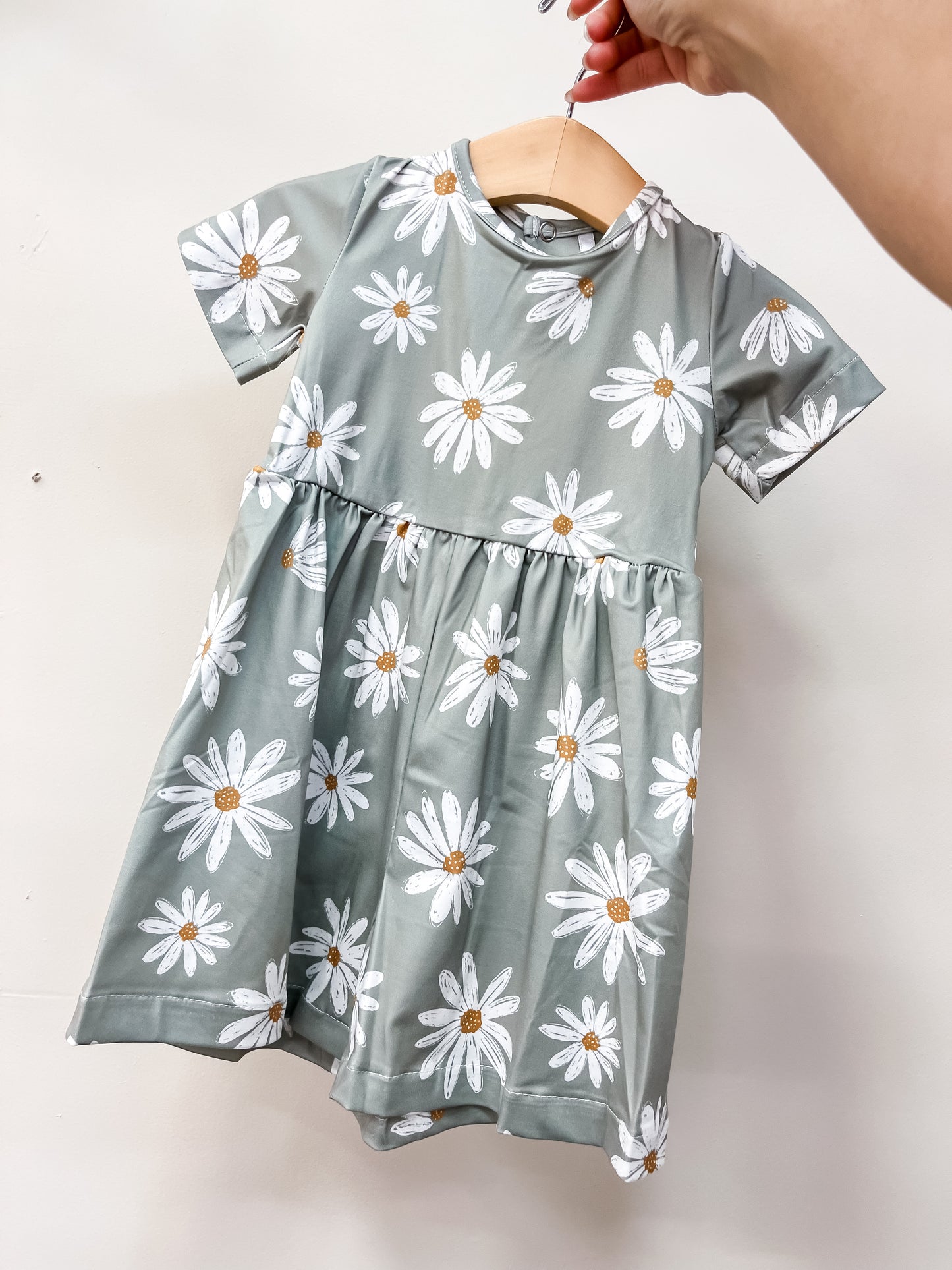 A hand holds up the Tiny Tot Bundles Daisy Daydream Romper—a short-sleeved, sage green children’s romper featuring a white daisy pattern and gathered waist—against a plain light background.