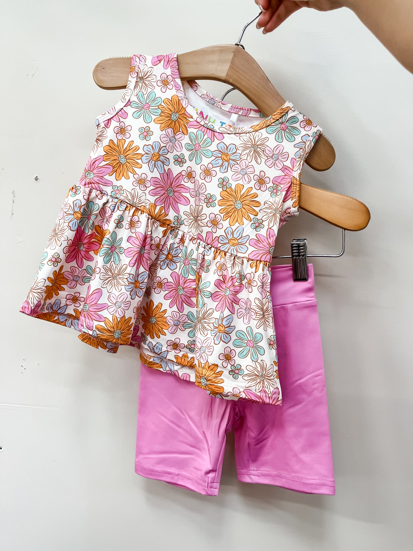 A hand holds a hanger displaying the Tiny Tot Bundles Petal Pop Peplum Set: a toddler outfit with a sleeveless floral top in pink, yellow, and white, paired with bright pink shorts, shown against a plain white background.