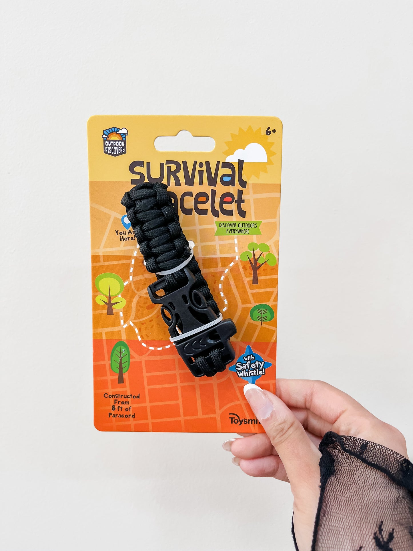 A hand holds a Toy Smith Discovery Survival Bracelet with Whistle in black. The orange package shows outdoor graphics, trees, and the text “Survival Bracelet” and “Constructed of 8 ft of paracord.”.