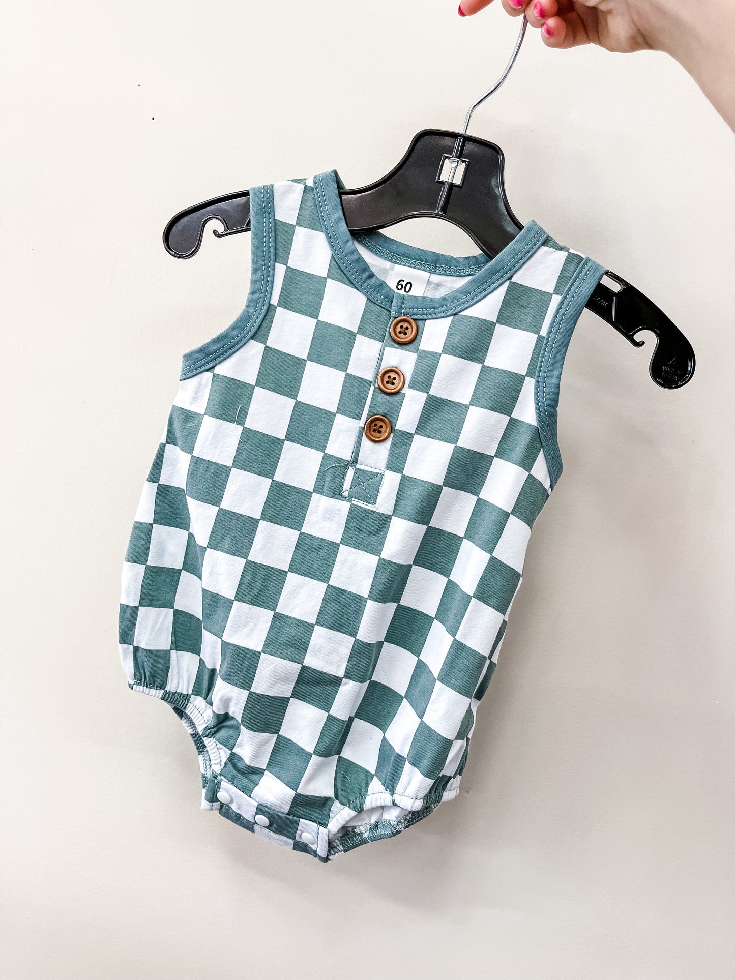 A hand holds a hanger displaying the Tiny Tot Bundles Checkmate Tank Romper, a sleeveless baby outfit with a green and white checkered pattern and three brown buttons on the front.
