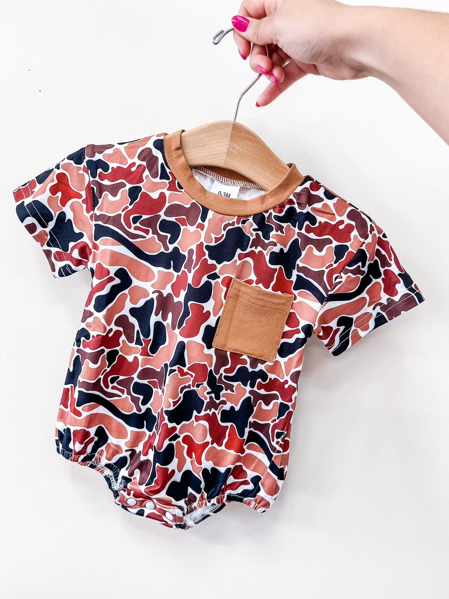 A hand with pink nails holds a Tiny Tot Bundles Rust Camo Bubble baby romper, featuring a brown, red, and black abstract camouflage pattern and a solid brown chest pocket.