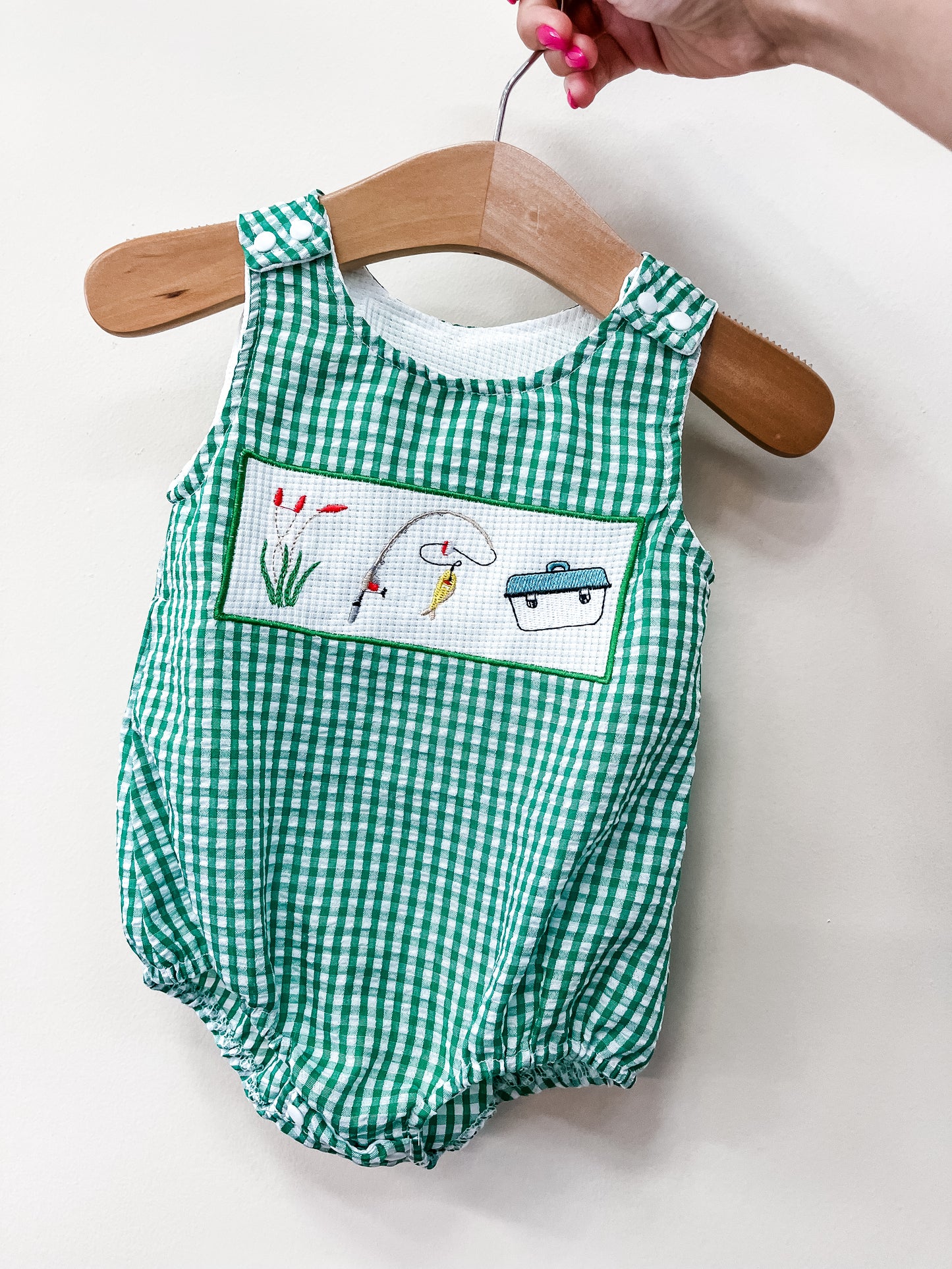 The Little Catch Bubble by Tiny Tot Bundles is a green and white gingham baby romper with fishing-themed embroidery on the front, displayed on a wooden hanger held by a hand with pink nail polish.