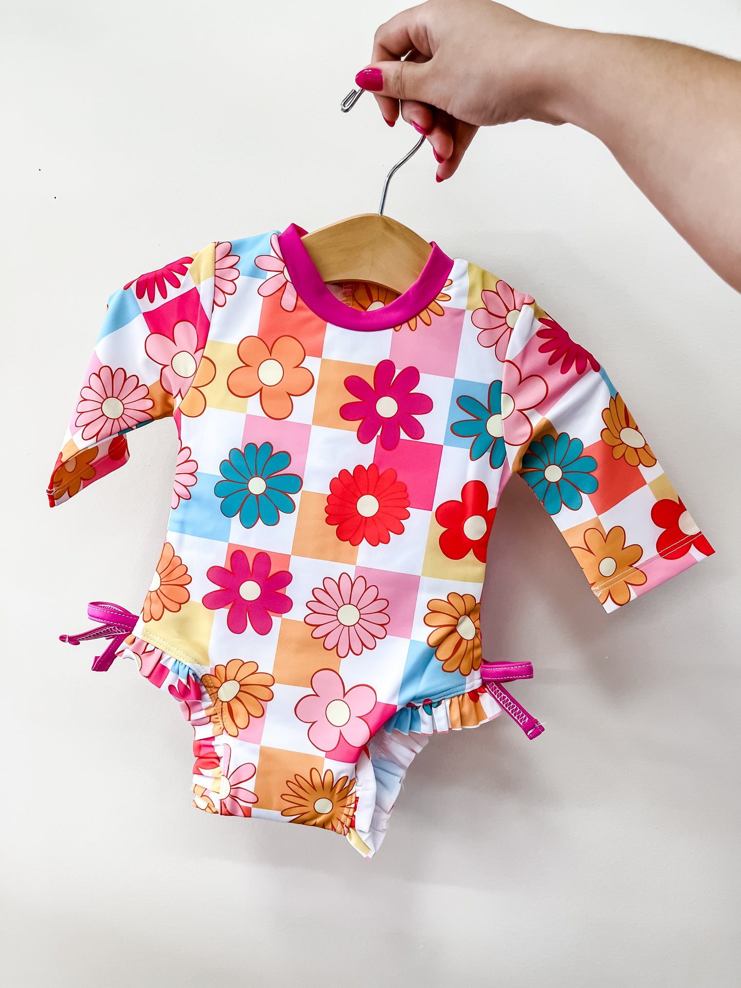 A hand holds a hanger with the Tiny Tot Bundles Groovy Garden Swimsuit, a long-sleeve baby suit featuring a pink, orange, and blue floral checkered pattern and pink trim, displayed against a plain white background.