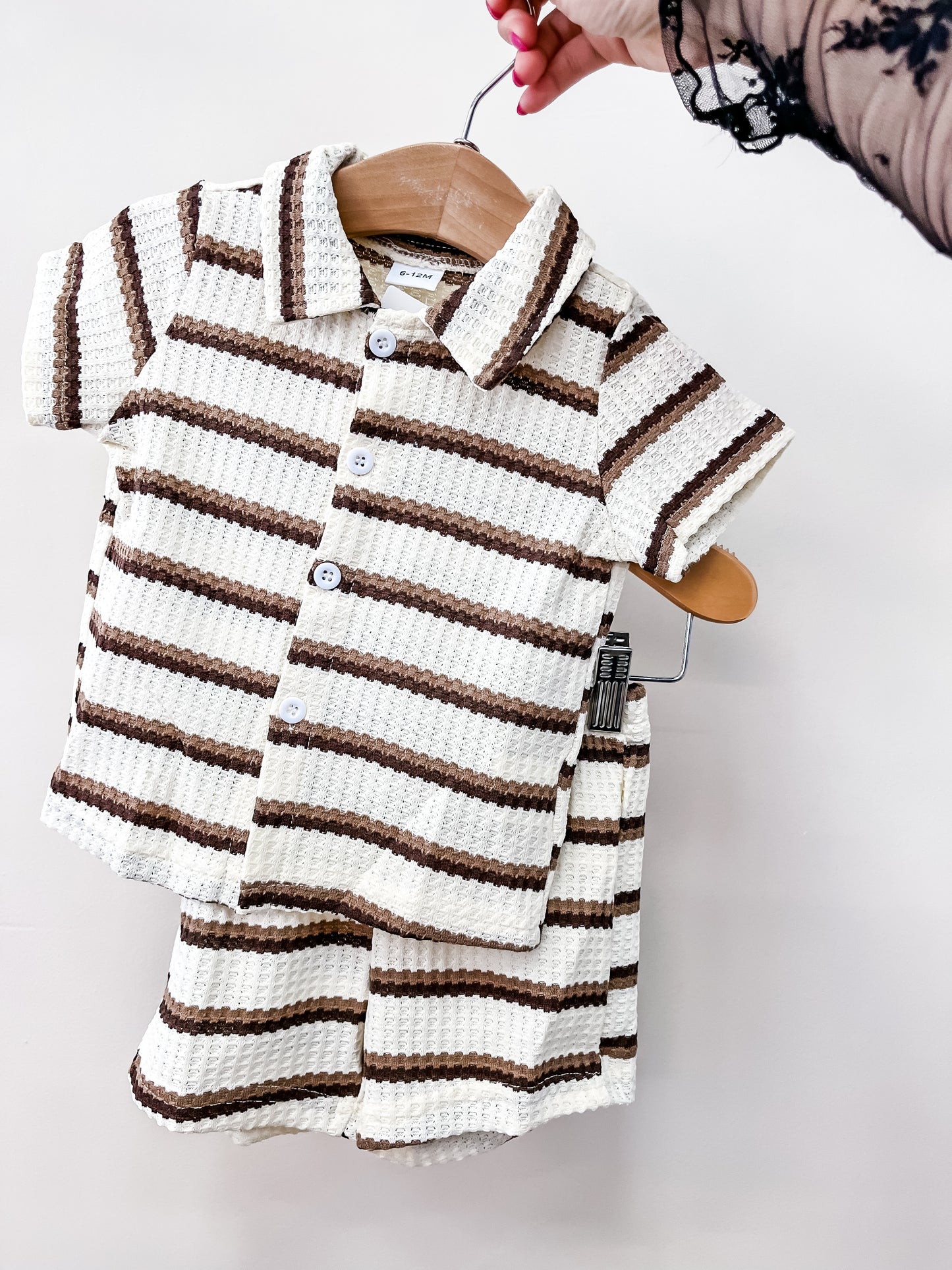 A hand holds the Mocha Coast Stripe Set by Tiny Tot Bundles, featuring a matching collared short-sleeve shirt and shorts in textured knit with cream and brown horizontal stripes, perfect for children.