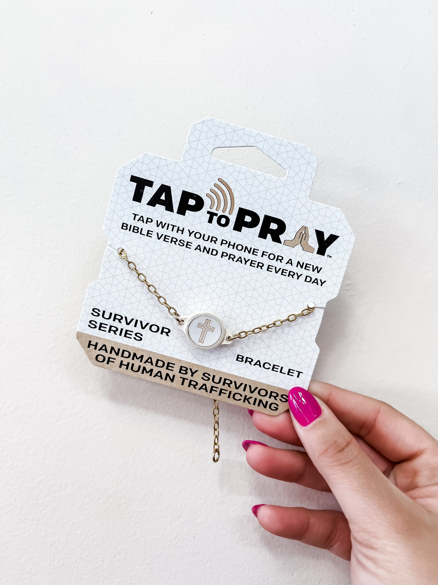 A hand with pink nails holds a versible Tap To Pray Bracelet - Gold Chain package, featuring a circular cross charm and noting it is handmade by survivors of human trafficking.