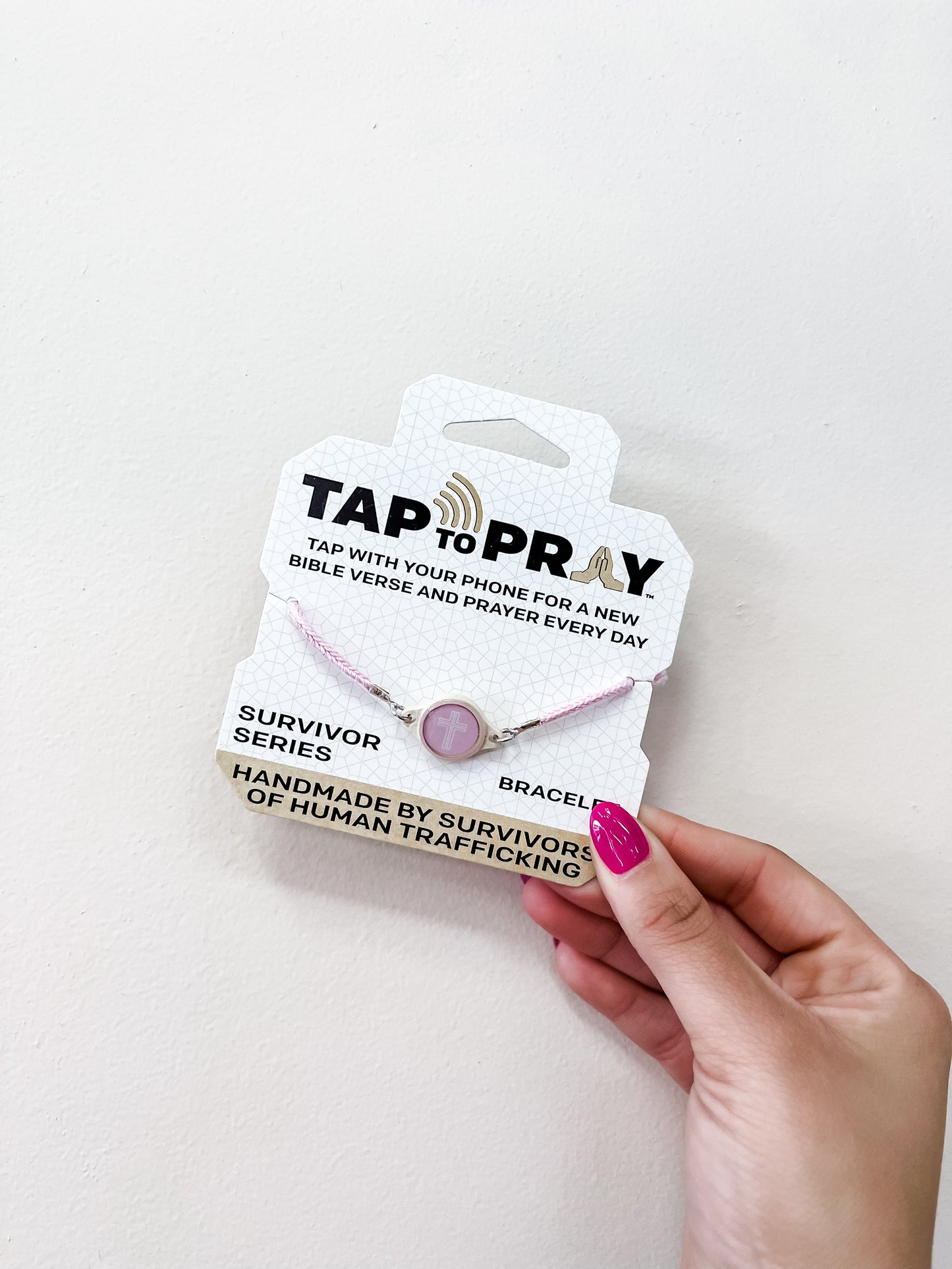 A hand with pink painted nails holds the versible Tap To Pray Bracelet - Lotus package, labeled handmade by survivors of human trafficking. The white packaging displays a pink bracelet with a circular charm.