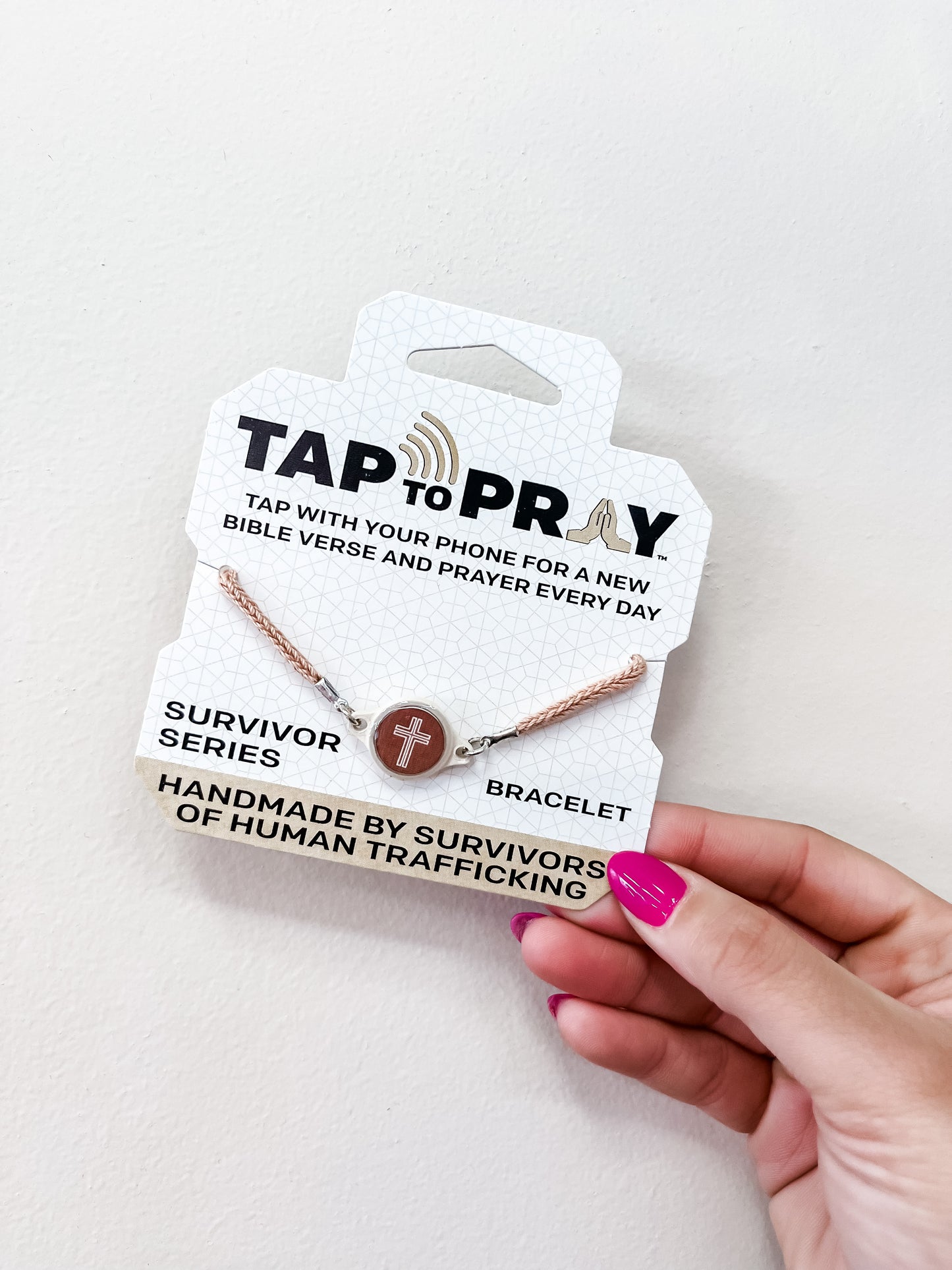 A hand with pink nail polish holds a versible "Tap To Pray Bracelet - Mocha" package. The bracelet features a cross charm and the packaging notes it is handmade by survivors of human trafficking.