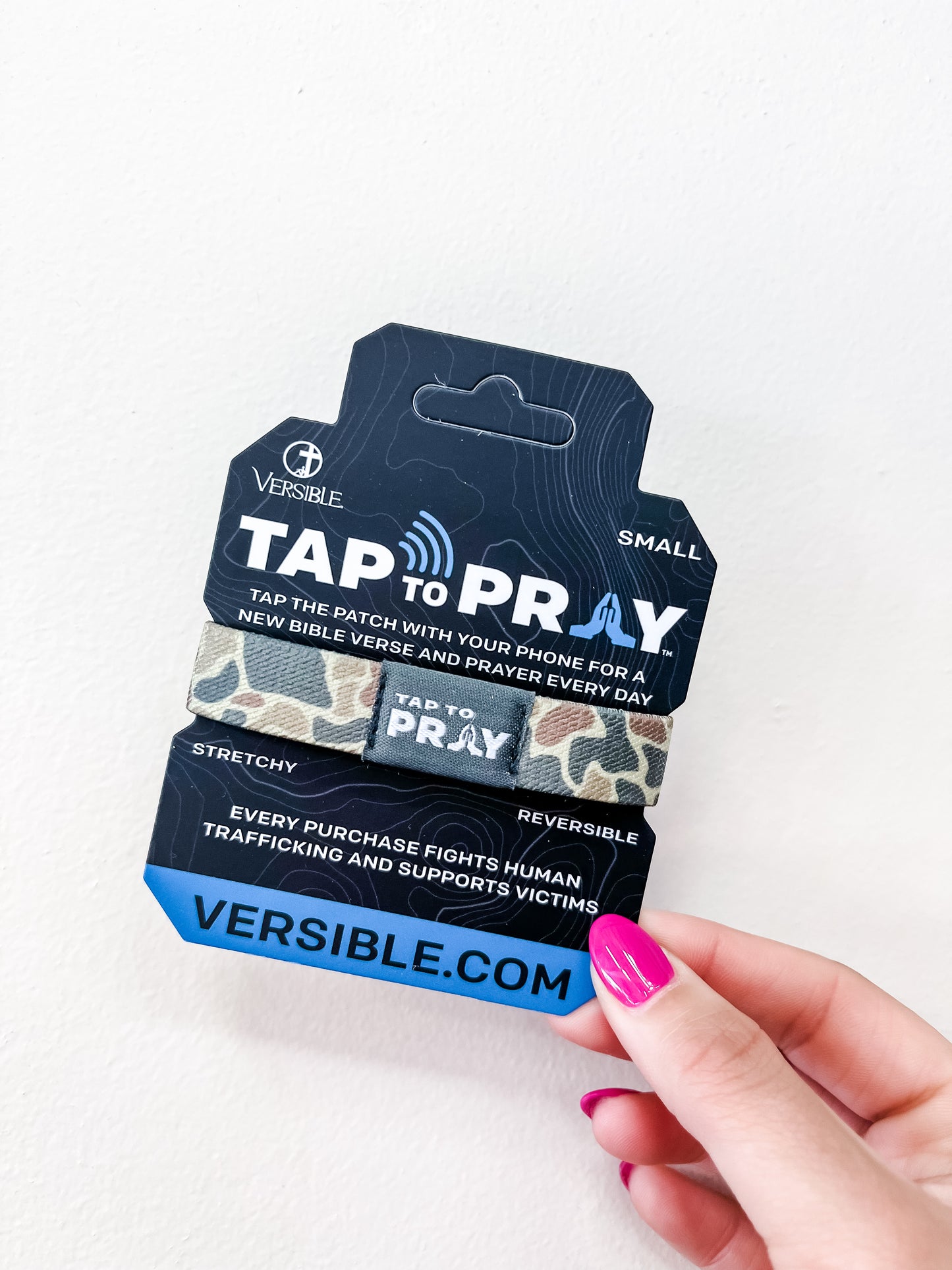 A hand with pink nail polish holds a versible Tap To Pray Bracelet - Sahara Camo package, supporting human trafficking victims and featuring the website VERSIBLE.COM.