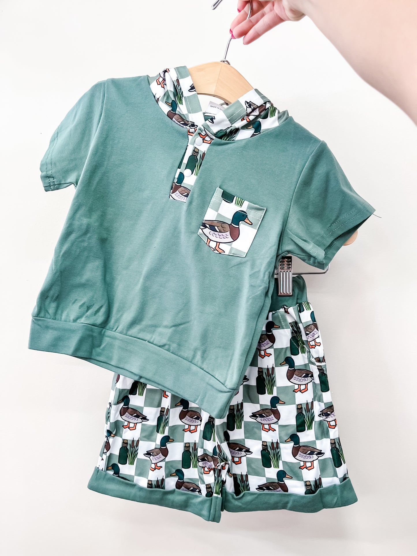 A hand holds up the Tiny Tot Bundles Pond Patrol Short Set— a green short-sleeve hooded shirt with a duck pocket print and matching shorts featuring an all-over duck and reeds pattern, displayed on a hanger against a white background.