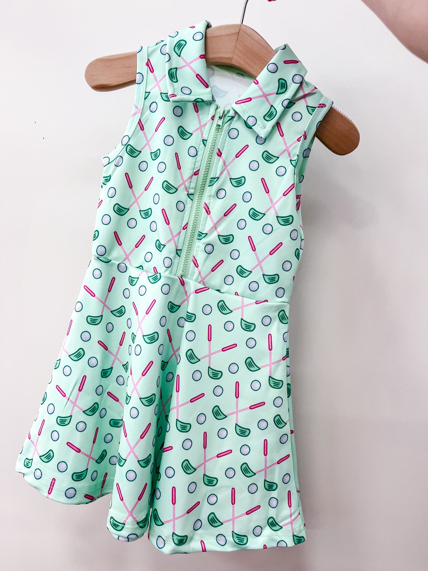 The Tiny Tot Bundles Green Quarter Zip Golf Dress with a collar, sleeveless cut, and pink hockey stick and green puck pattern hangs on a wooden hanger against a plain light background.