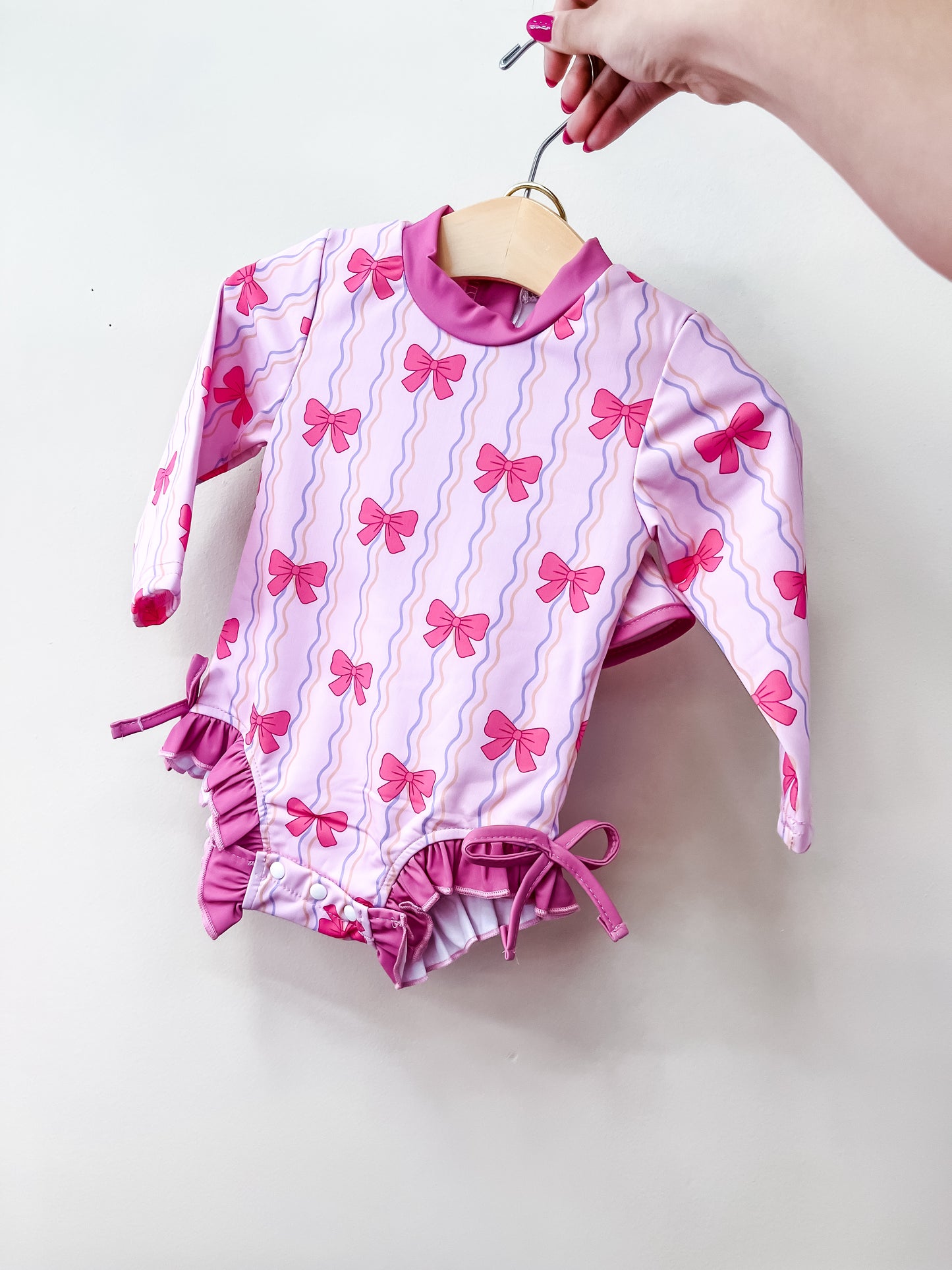 A hand holds the Tiny Tot Bundles Pink Bow Swimsuit, a long-sleeved baby swimsuit in pink and purple with bow and wavy line patterns, against a plain white background.