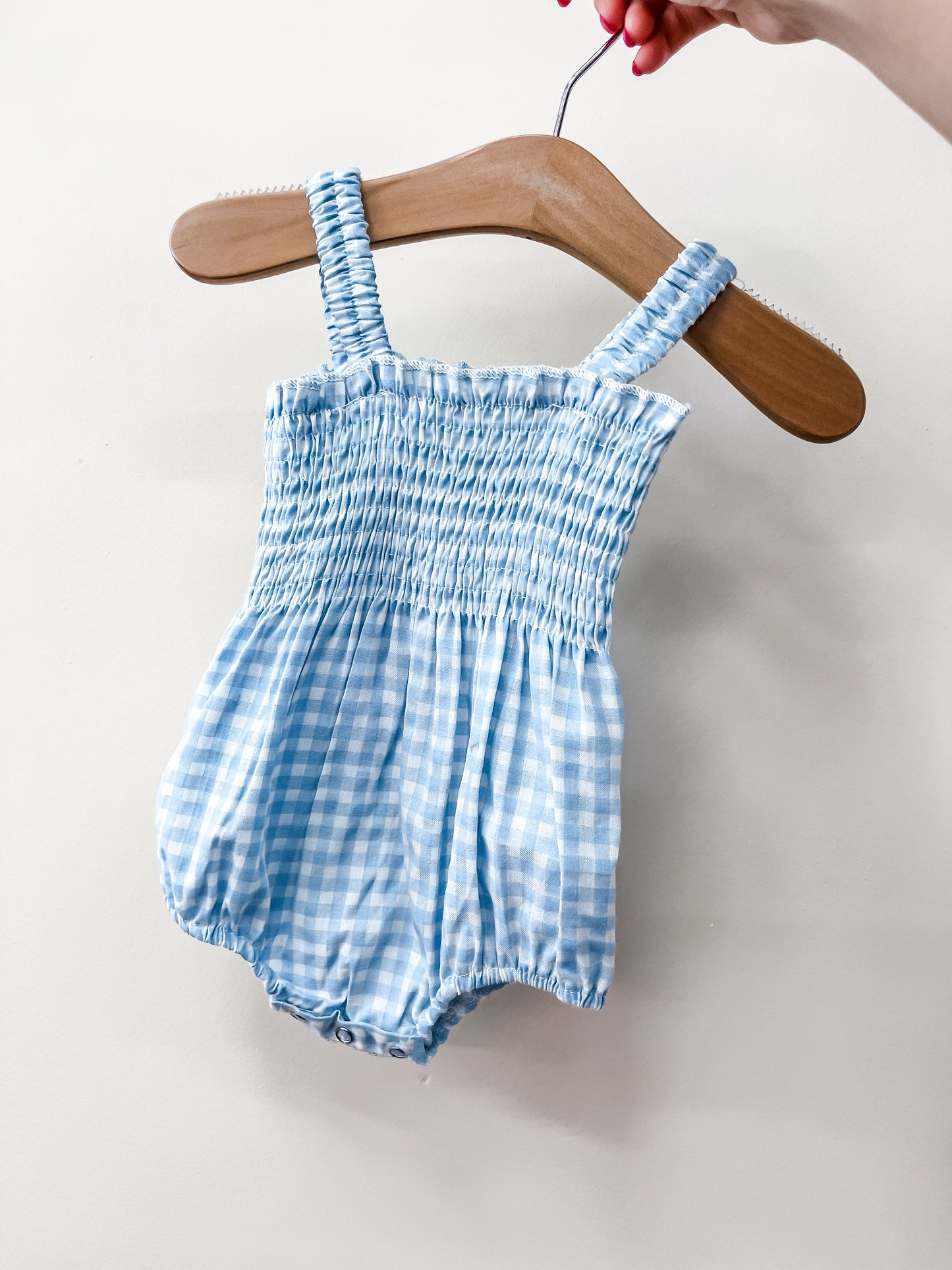 A hand holds the Tiny Tot Bundles Sweet Sky Smocked Romper, featuring blue and white gingham fabric with shirred elastic straps and bodice, displayed on a wooden hanger against a plain white background.