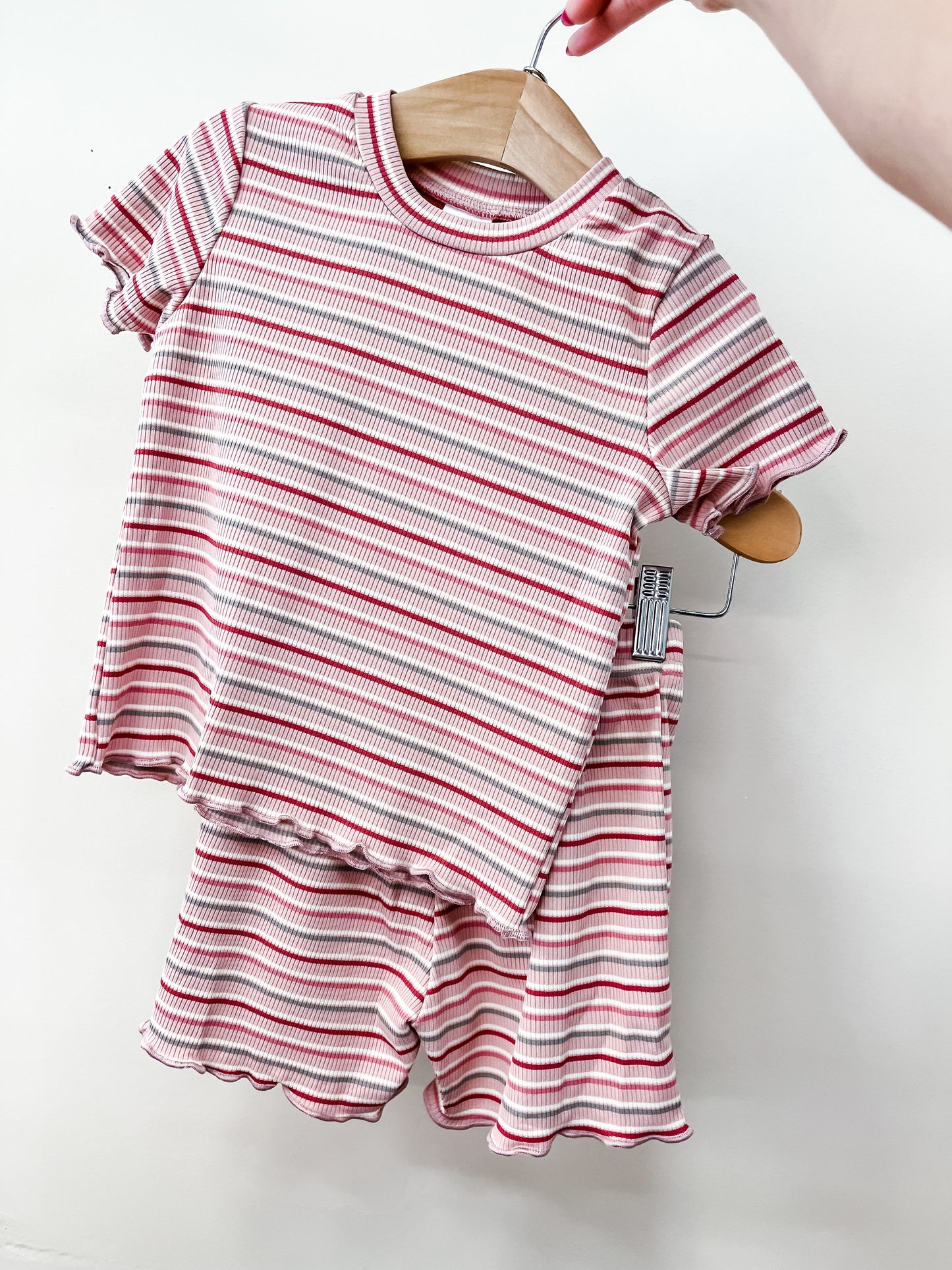 A hand holds a Tiny Tot Bundles Rosy Stripe Snuggle Set—a matching children's outfit with a pink and white striped, short-sleeved top and ruffled shorts—on a wooden hanger against a plain white background.